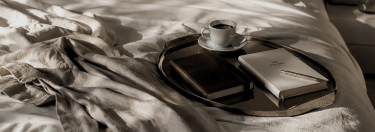 Tray with a cup of coffee, a closed book, a planner, and a notepad on a bed with rumpled sheets.