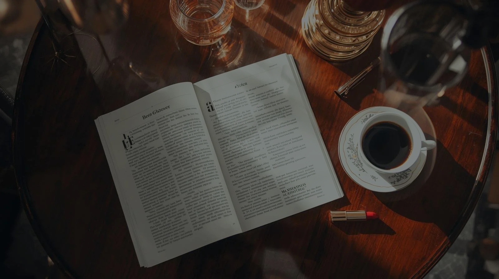 A wooden table with an open magazine, a cup of black coffee on a decorative saucer, a lipstick, and glassware, with some sunlight casting shadows.