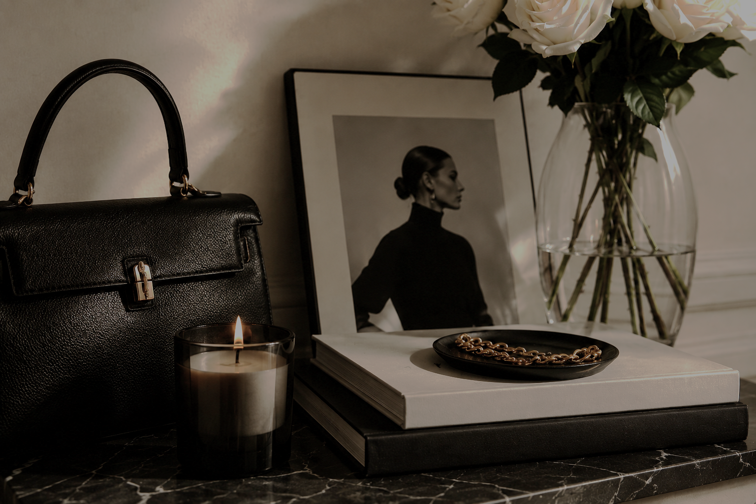 A tabletop display with a black textured handbag, a lit candle in a dark glass holder, an open book with a black cover, a black plate with a gold chain, a framed black and white portrait of a woman, and a clear glass vase with white roses.