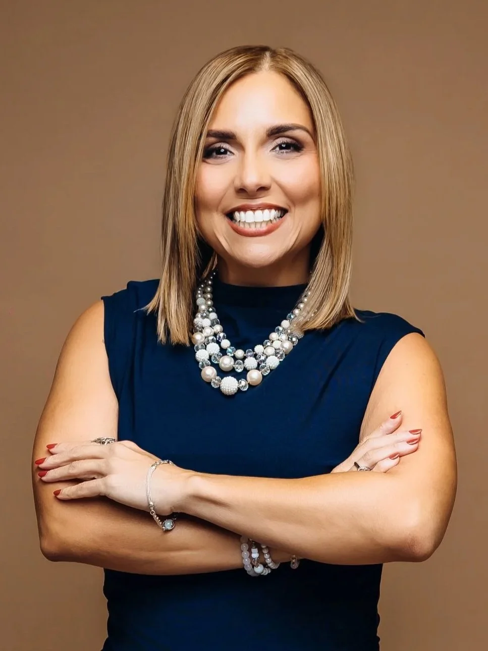 Dr. Evelyn M. Ortiz Robert, founder of Synergy Dots, smiling with arms crossed against a beige background. She has shoulder-length blonde hair and wears a navy blue sleeveless top with multiple pearl necklaces.