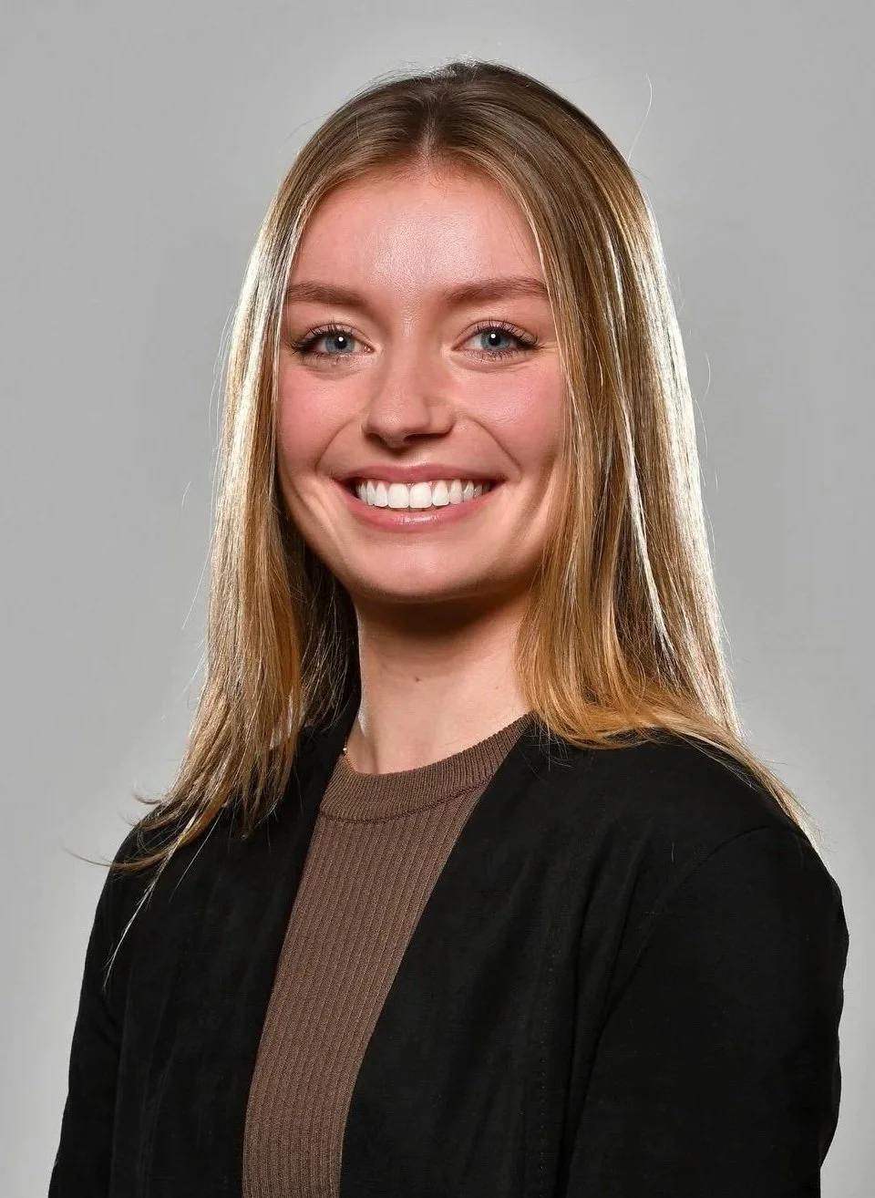A young woman with long blonde hair smiling at the camera, wearing a brown top and a black jacket, against a gray background.