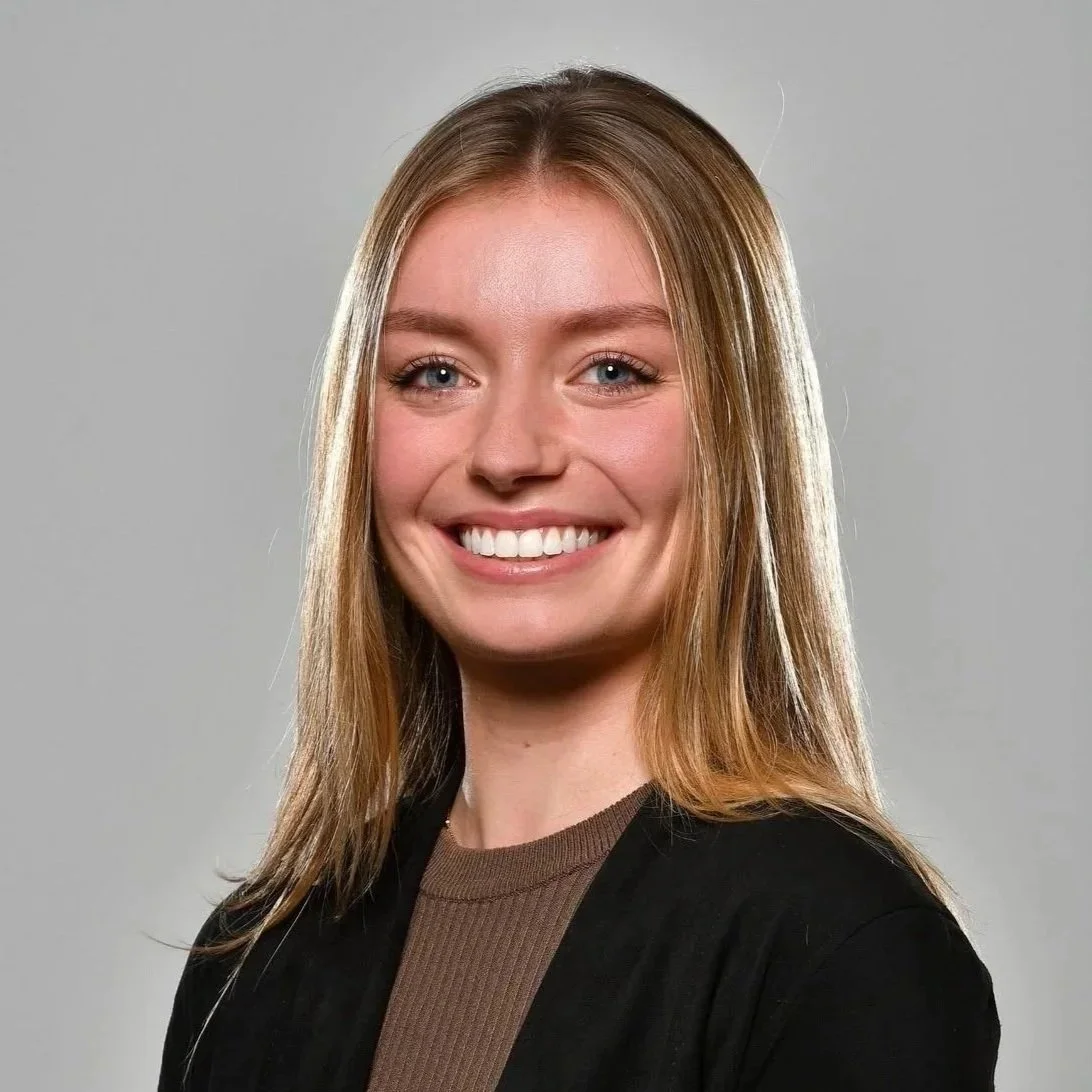 A woman with long, straight, light brown hair smiling with teeth showing, wearing a brown top and black jacket, against a plain gray background.