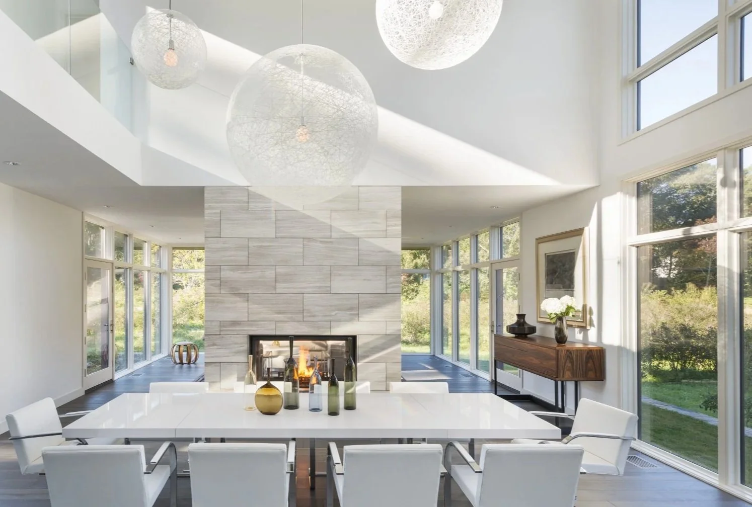 Modern dining room with large windows, white dining table, white chairs, a fireplace, decorative vases, and a wooden sideboard, illuminated by natural light.