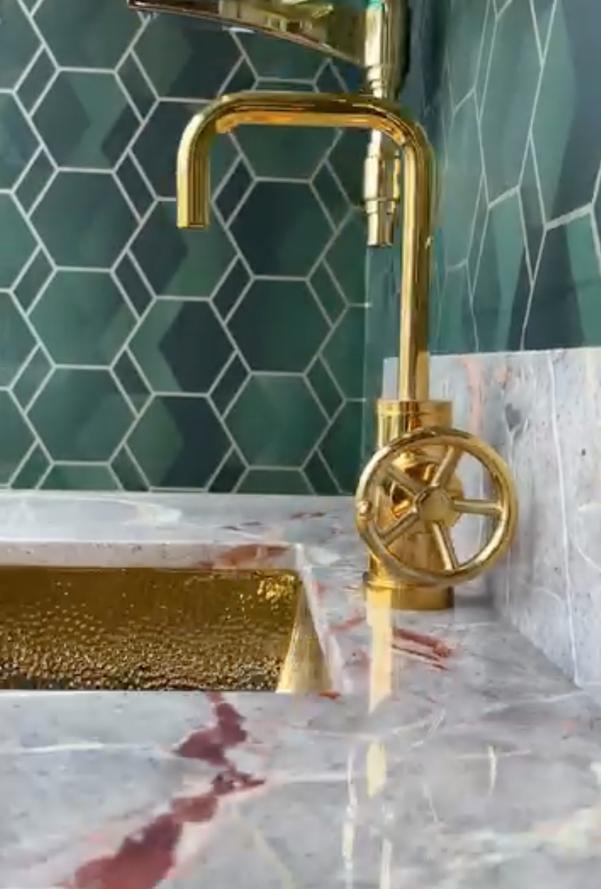 Gold kitchen faucet with a gold handle on a marble countertop, with a green hexagonal tile backsplash.