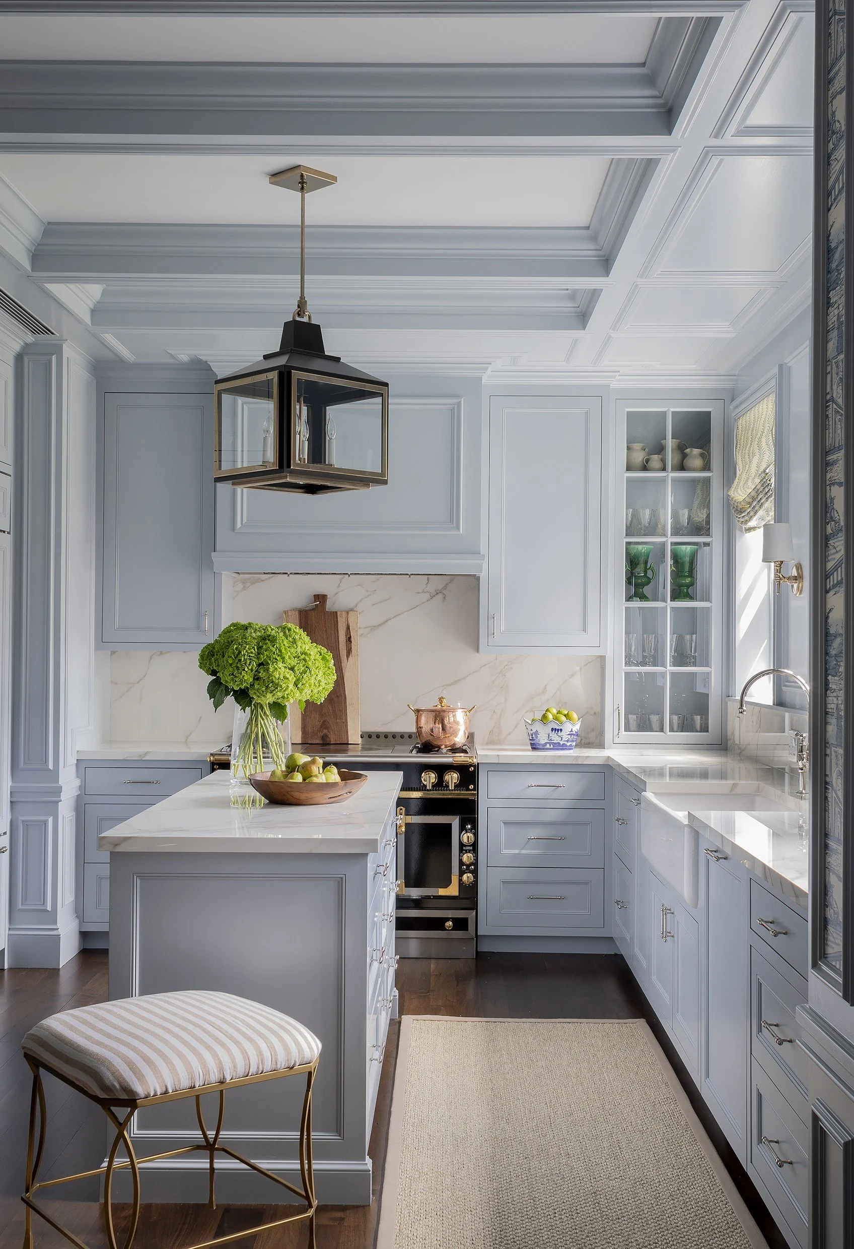 A modern kitchen with light blue cabinets, a white marble countertop, a kitchen island with a marble top, a small round stool with a striped beige cushion, a green hydrangea plant in a glass vase, a copper teapot, a blue and white patterned bowl with green apples, a built-in glass cabinet with green and clear glassware, a window with a patterned curtain, and a pendant light hanging from the ceiling.