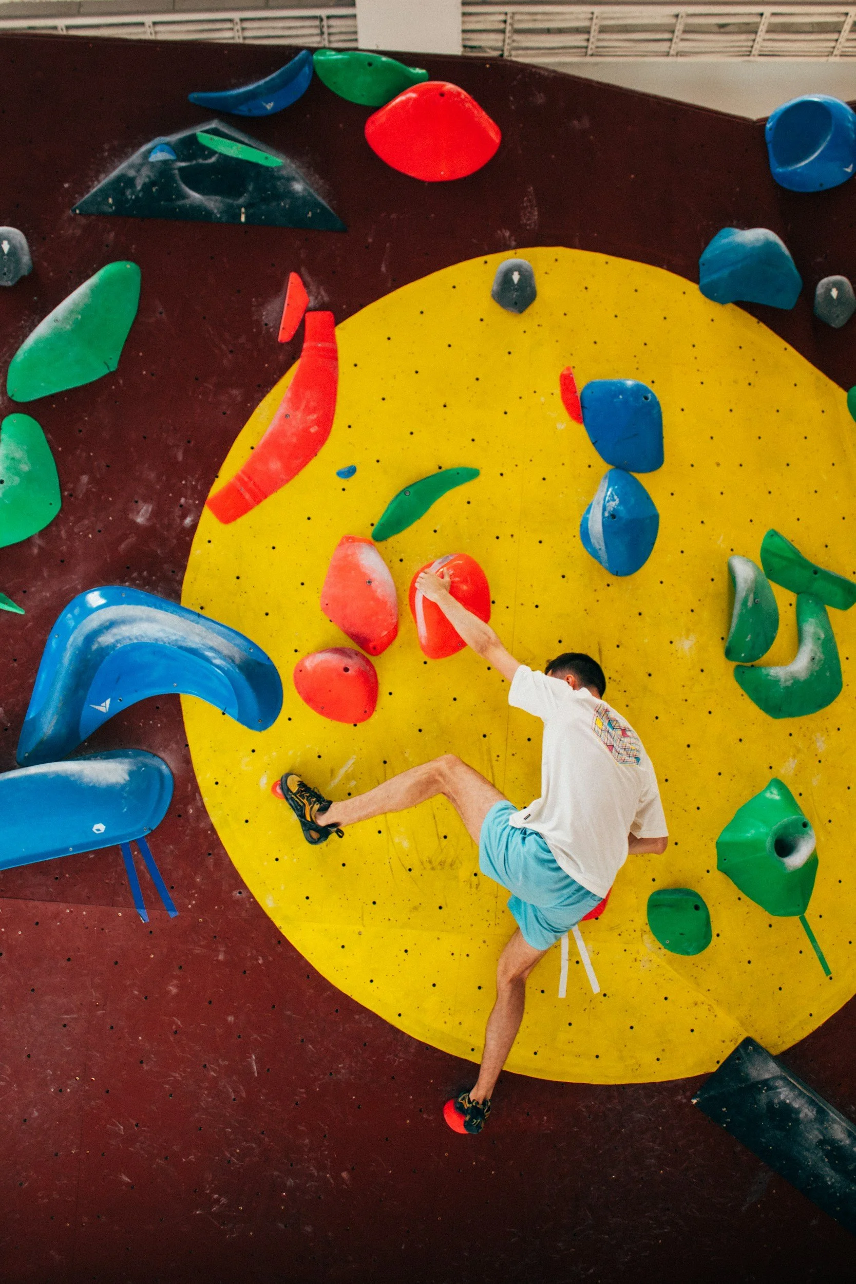 A person wearing a white shirt, light blue shorts, and climbing shoes is climbing an indoor bouldering wall with various colorful holds, including red, green, blue, and black.