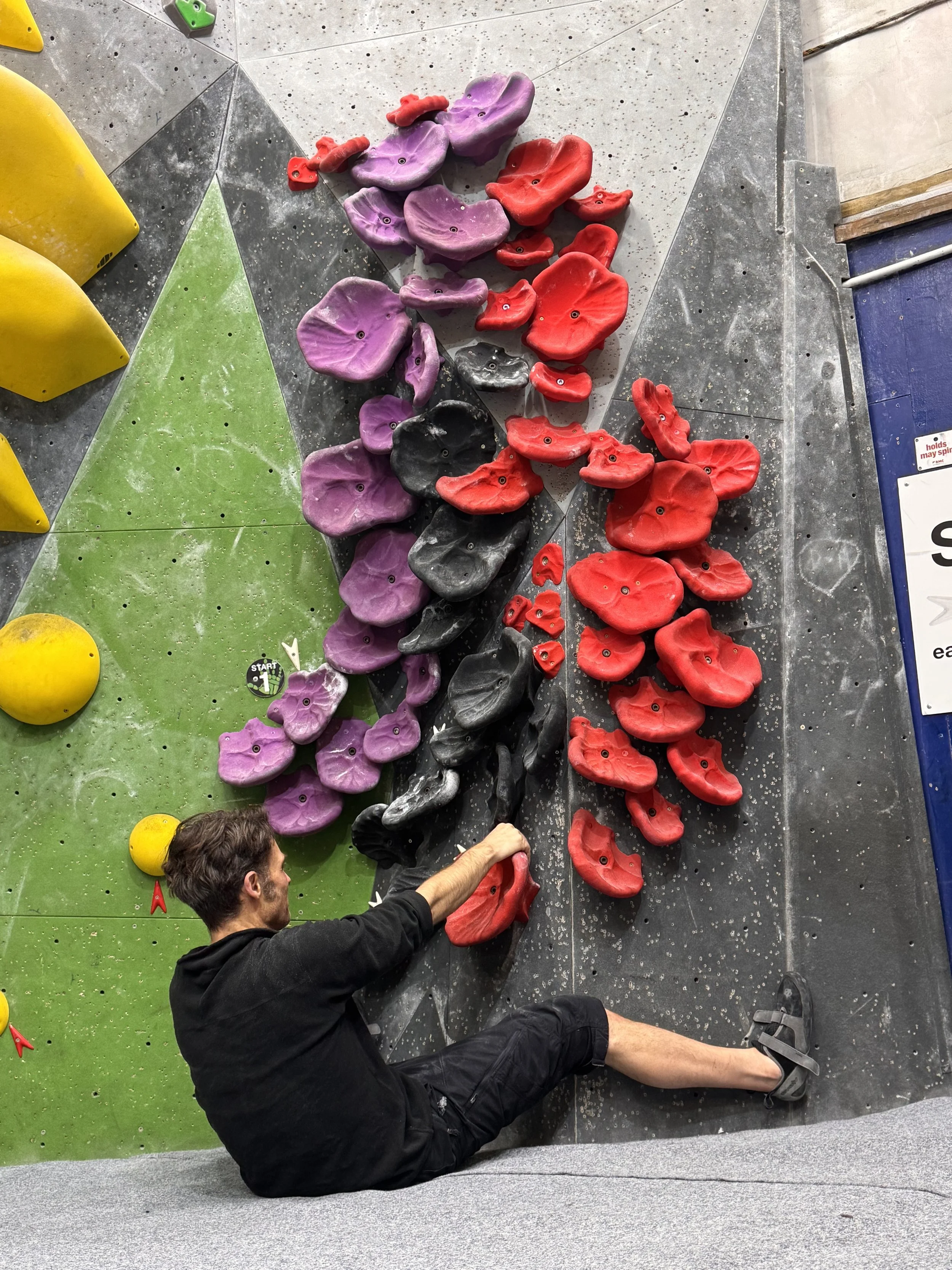 A man sitting on the floor in front of an indoor rock climbing wall with colorful holds, including purple, red, black, and yellow, extending a hand towards the holds. His face is partially visible, and he is wearing black clothing and climbing shoes.