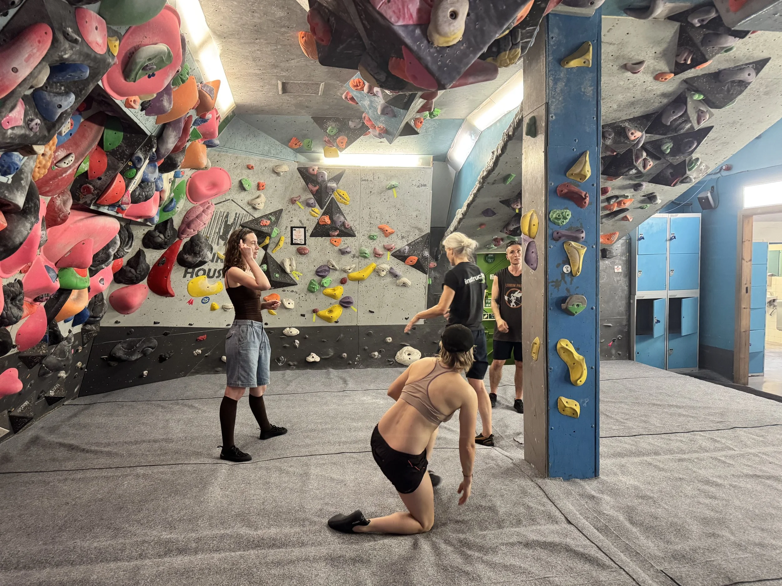 Indoor bouldering gym with climbing walls covered in various colorful holds and people practicing climbing skills, some assisting others or resting.
