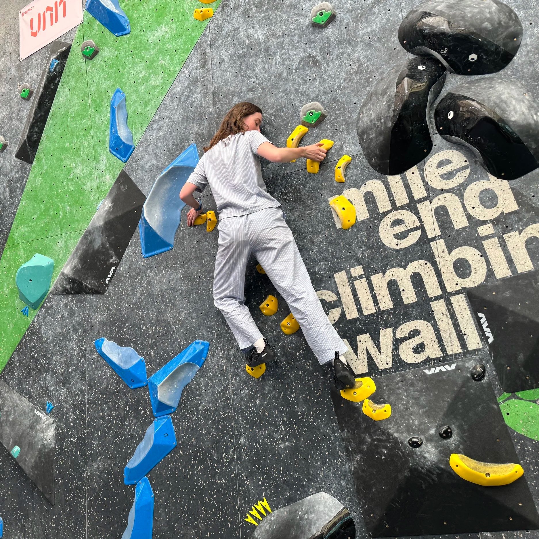 An indoor rock climbing gym with a woman in pajamas climbing on a wall with colorful holds, featuring green, yellow, blue, and black shapes, and black wall sections with white lettering.
