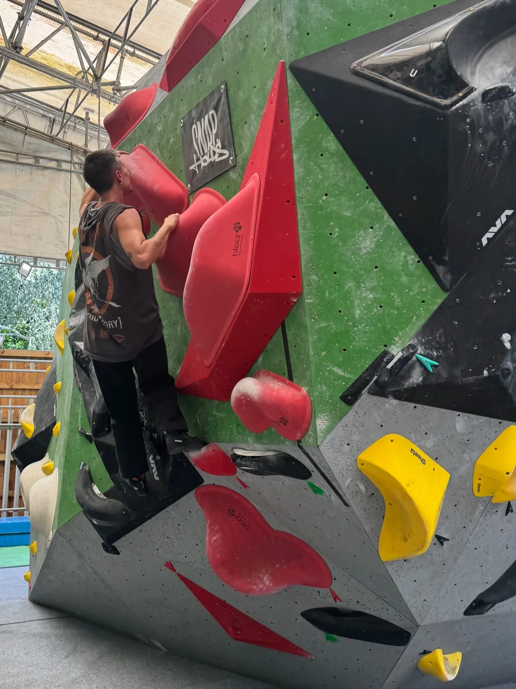 An indoor rock climbing gym with a person climbing a bouldering wall with large red holds and yellow holds, surrounded by a padded flooring.
