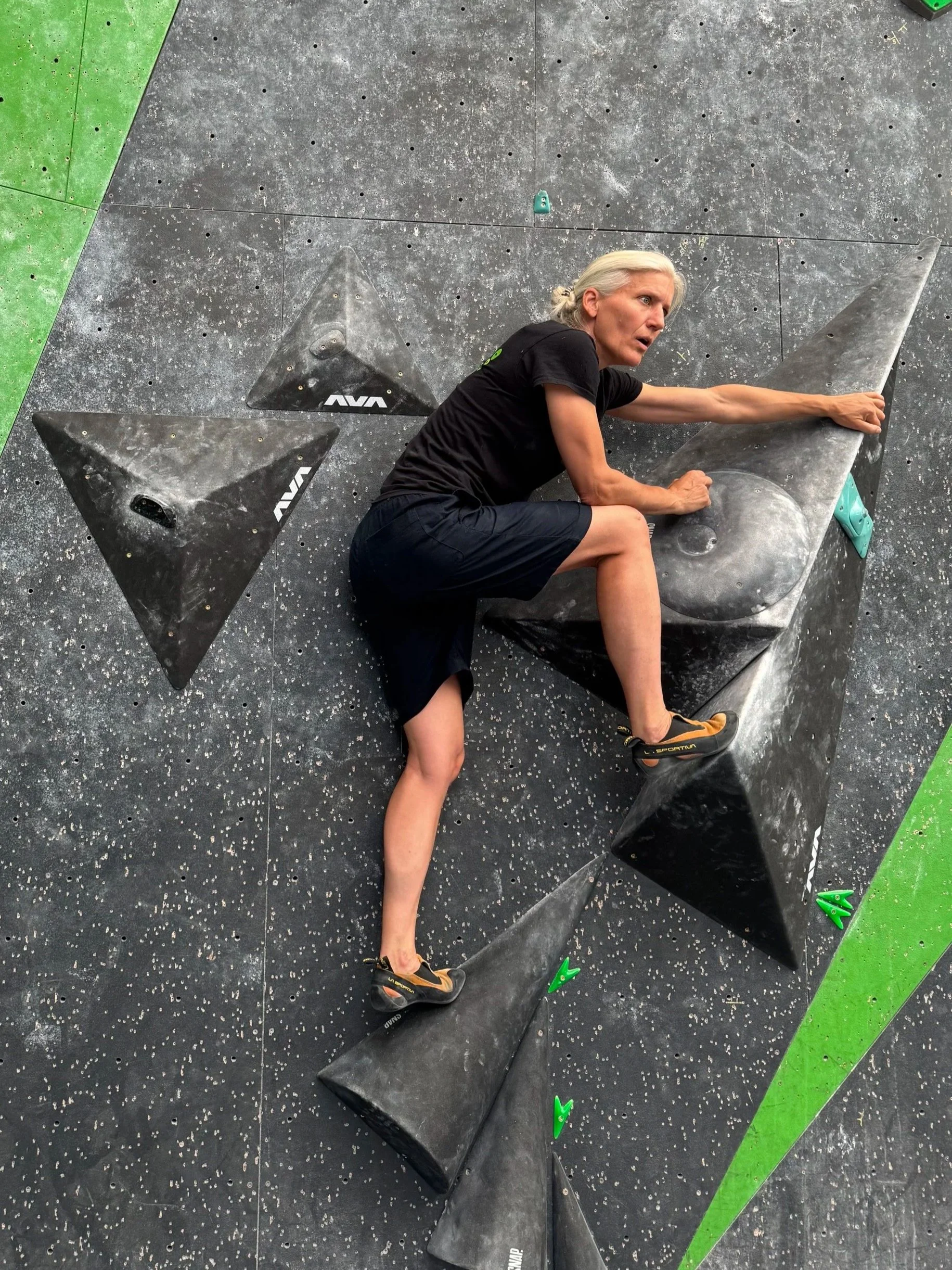 A woman with blonde hair in a bun is climbing an indoor bouldering wall, reaching for a hold with her right hand while her left hand grips a hold. She is wearing a black T-shirt, black shorts, and climbing shoes.