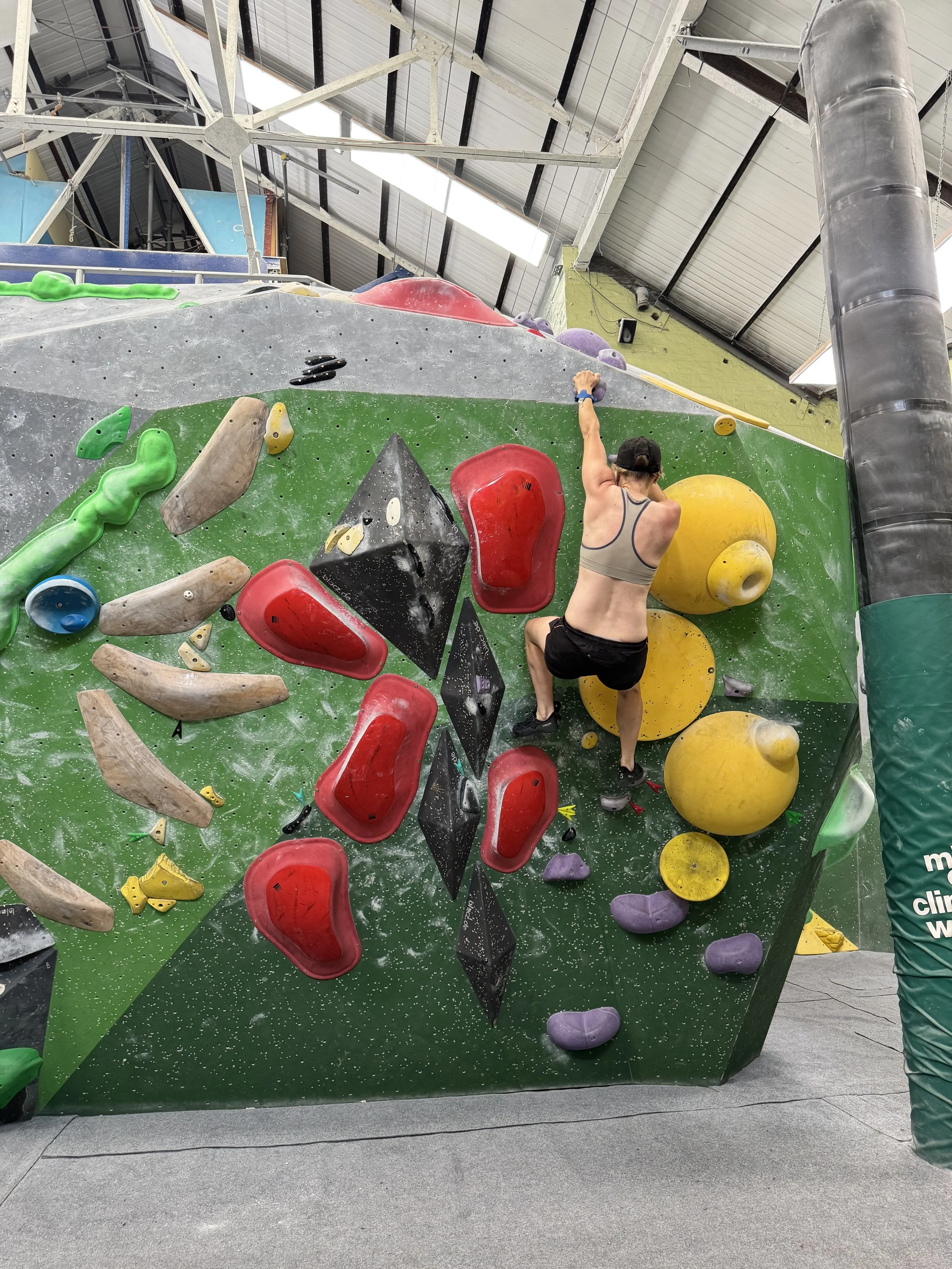 An indoor bouldering gym with a person climbing on a colorful climbing wall that has various holds and volumes. The gym has a high ceiling with visible metal beams and a ventilation duct.