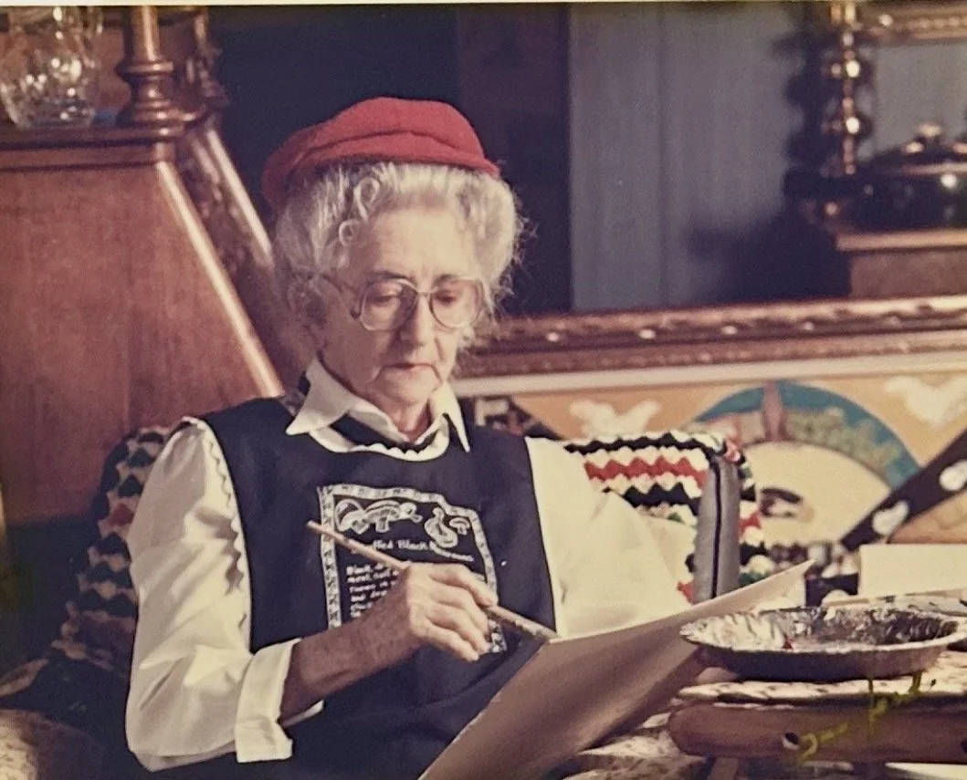 An elderly woman with gray curly hair, glasses, wearing a red hat and a black vest over a white shirt, is sitting on a colorful, patterned sofa and painting on a large sheet of paper.