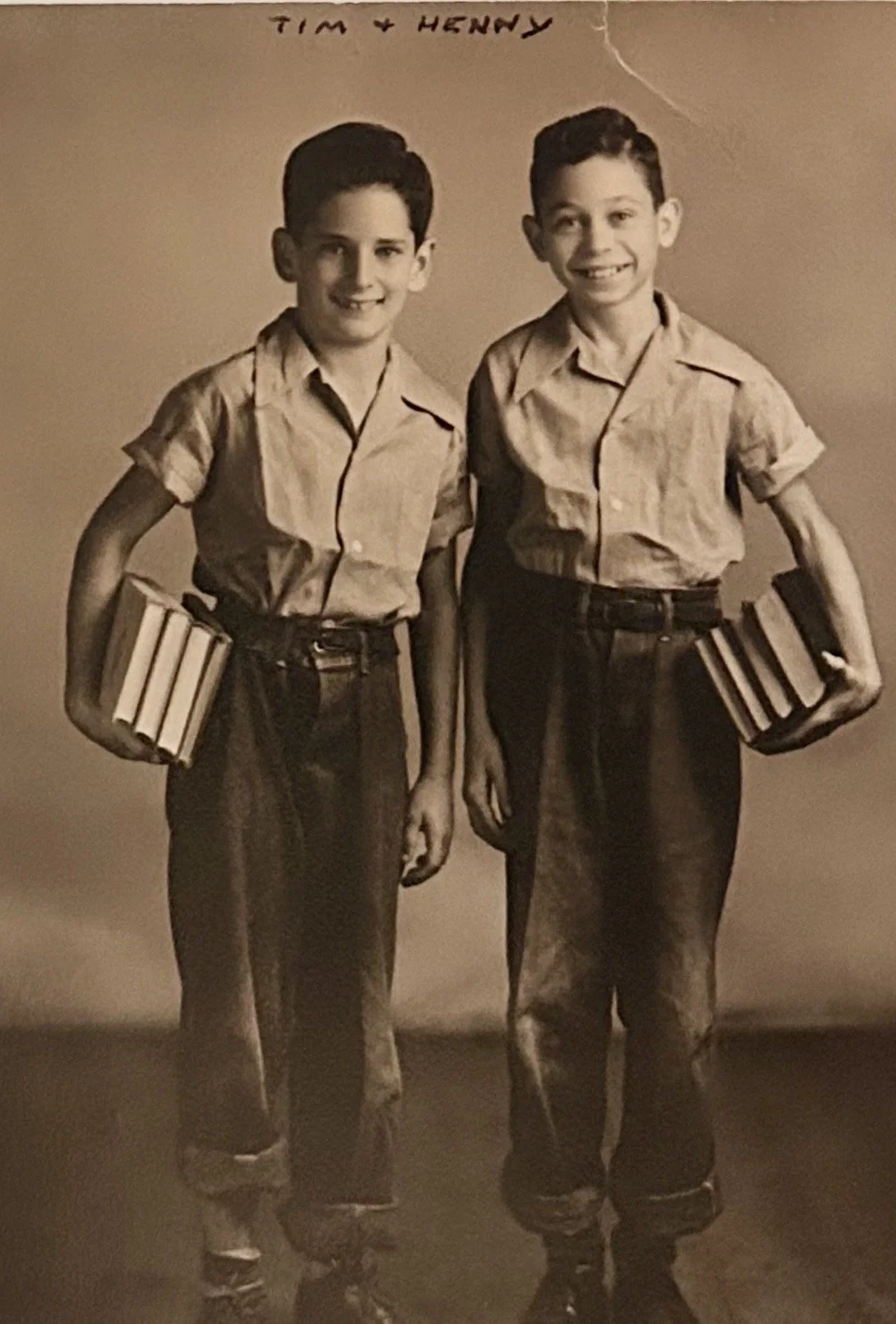 Black and white photo of two young boys standing side by side, both holding books and wearing similar short-sleeved shirts and dark pants. The names 'Tim' and 'Henry' are handwritten at the top of the photo.