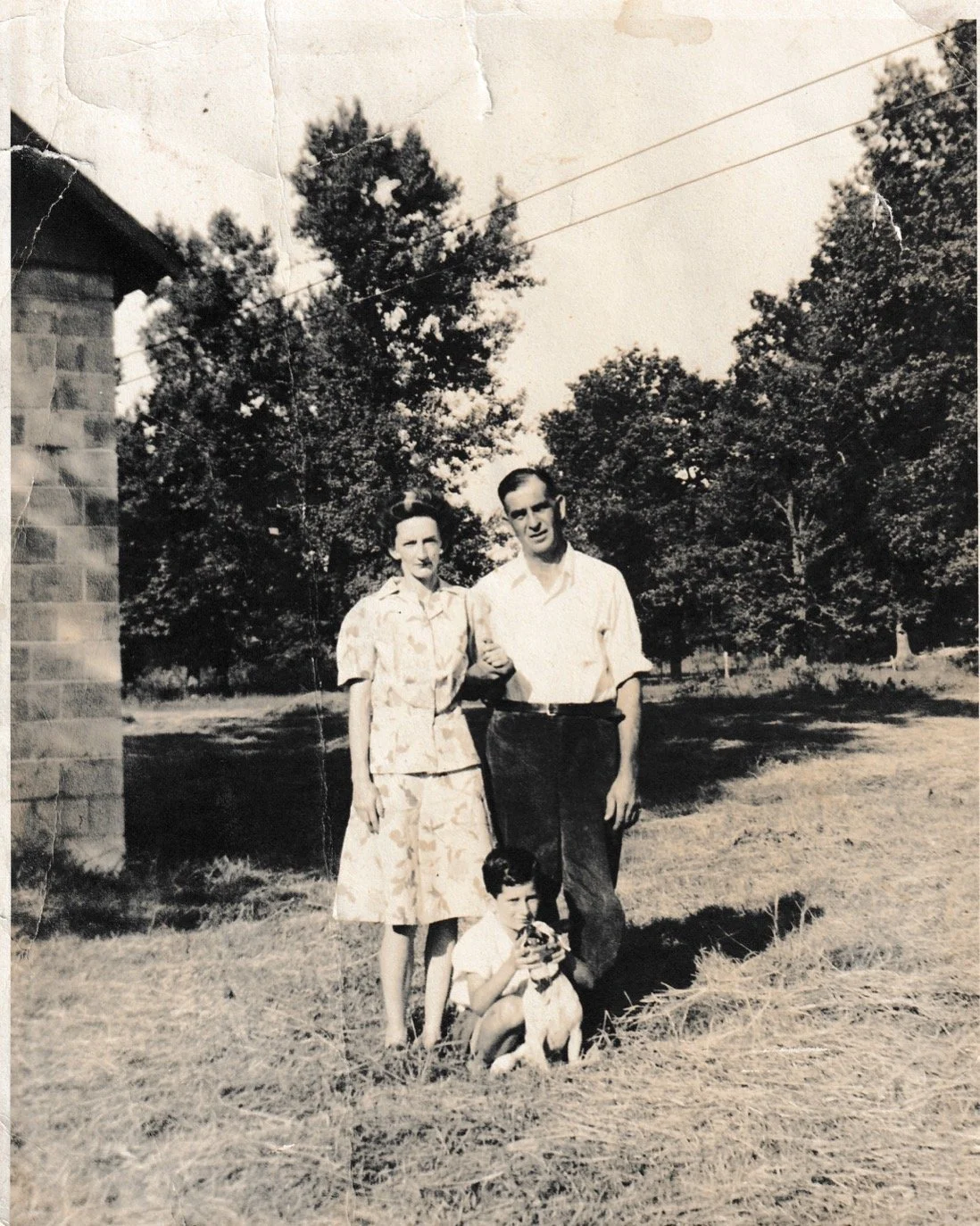 A black-and-white photograph of a family outdoors, consisting of a woman, a man, and a young boy seated on the ground with a dog. The background features trees and a building on the left.