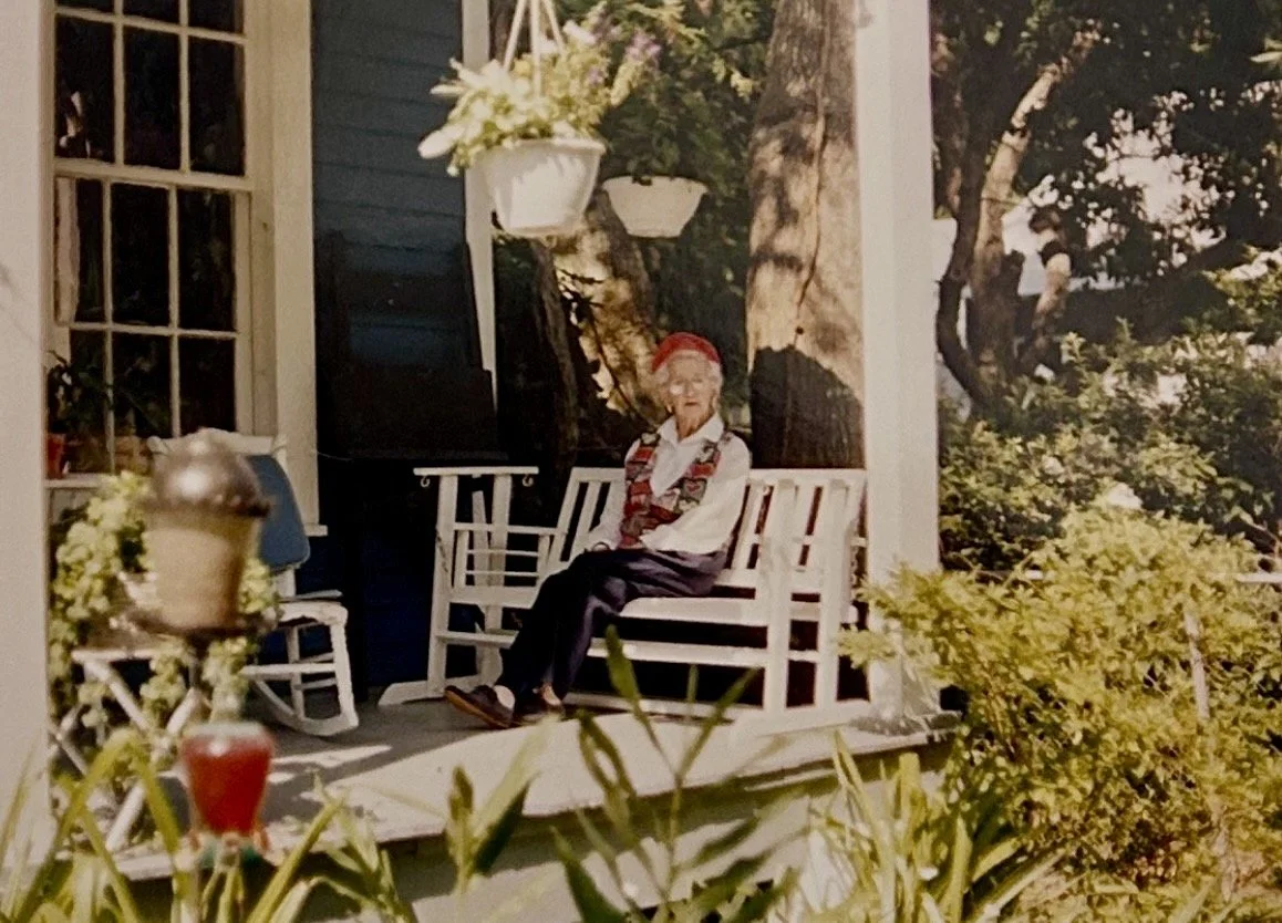 An elderly woman with a red headband sitting on a porch swing surrounded by plants and hanging flower baskets.