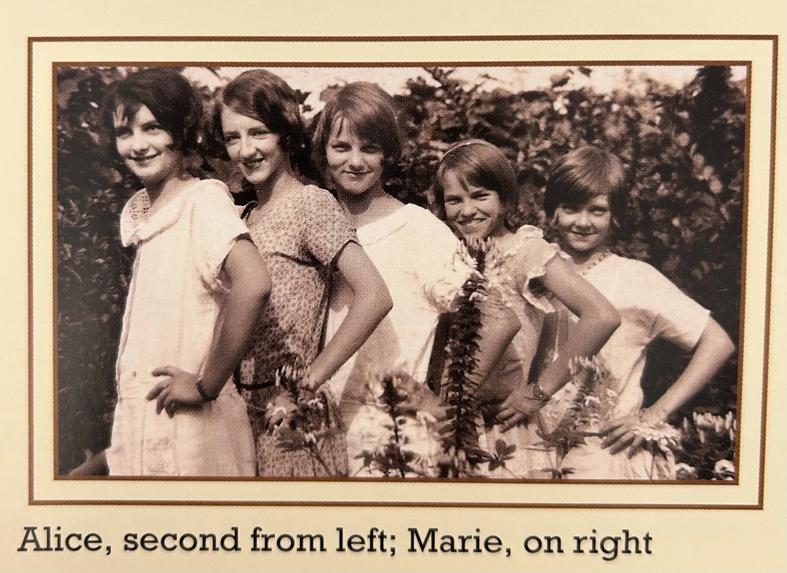 A black and white photograph of five young girls standing outdoors, smiling, with trees and foliage in the background. The caption indicates their names and positions.