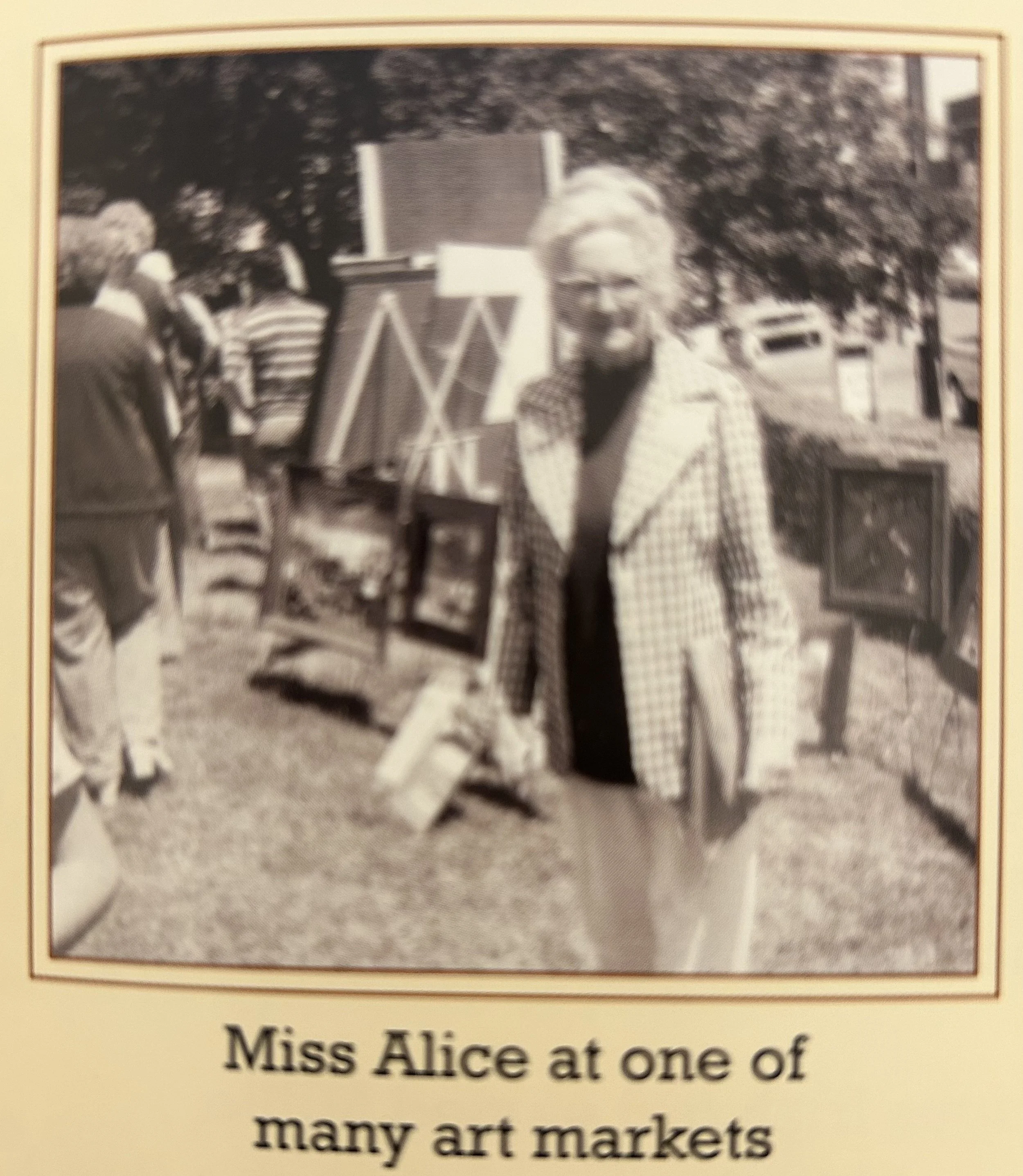 Black and white photograph of Miss Alice at one of many art markets, standing in front of her artwork with several people in the background.