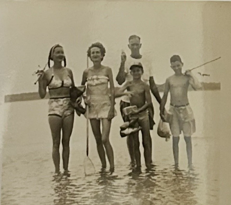 A black-and-white photo of five children standing in shallow water at the beach, smiling and holding fish and fishing rods.