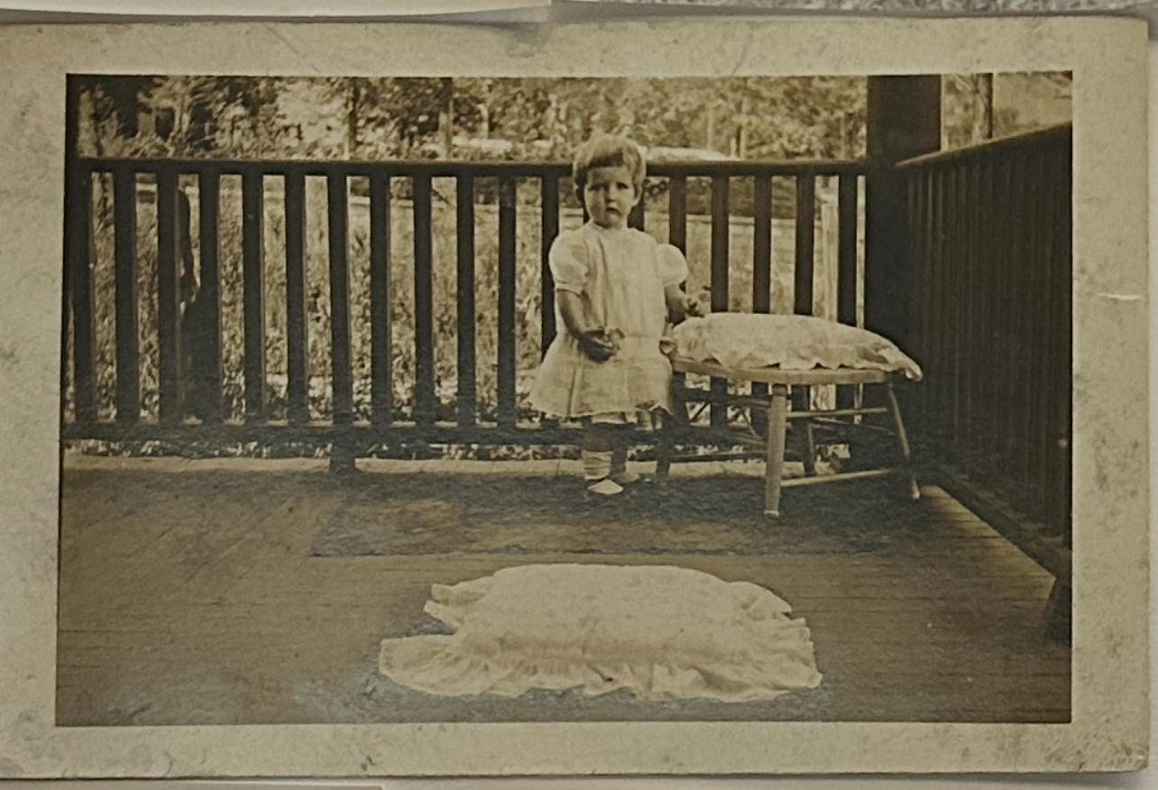 A young girl standing on a porch next to a small table with a pillow, with a blanket laid on the floor in front of her, outside a house with wooden railing and greenery in the background.