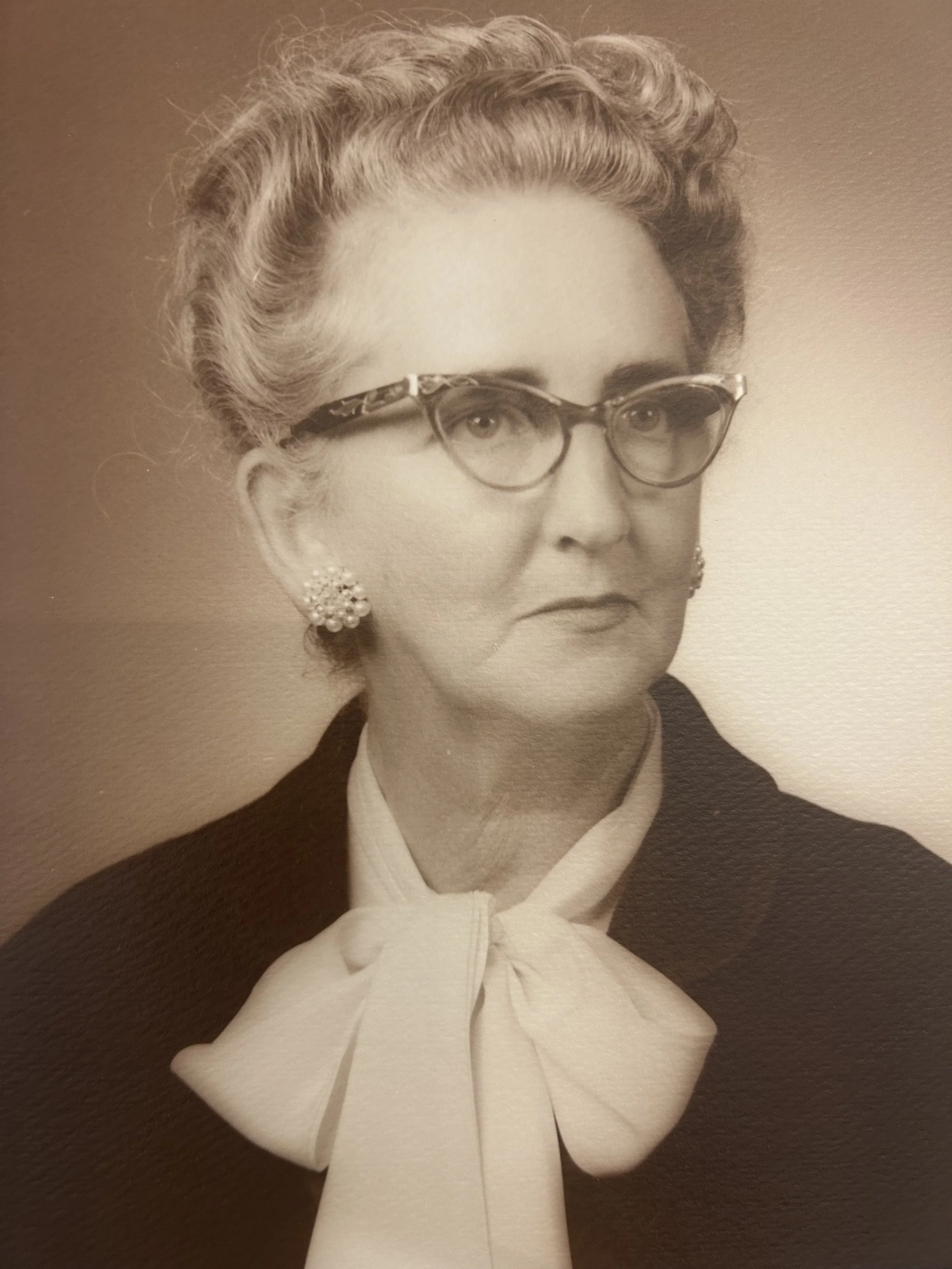 Black-and-white portrait of a woman with short, wavy hair, wearing glasses, pearl earrings, a dark jacket, and a white blouse with a bow at the collar.
