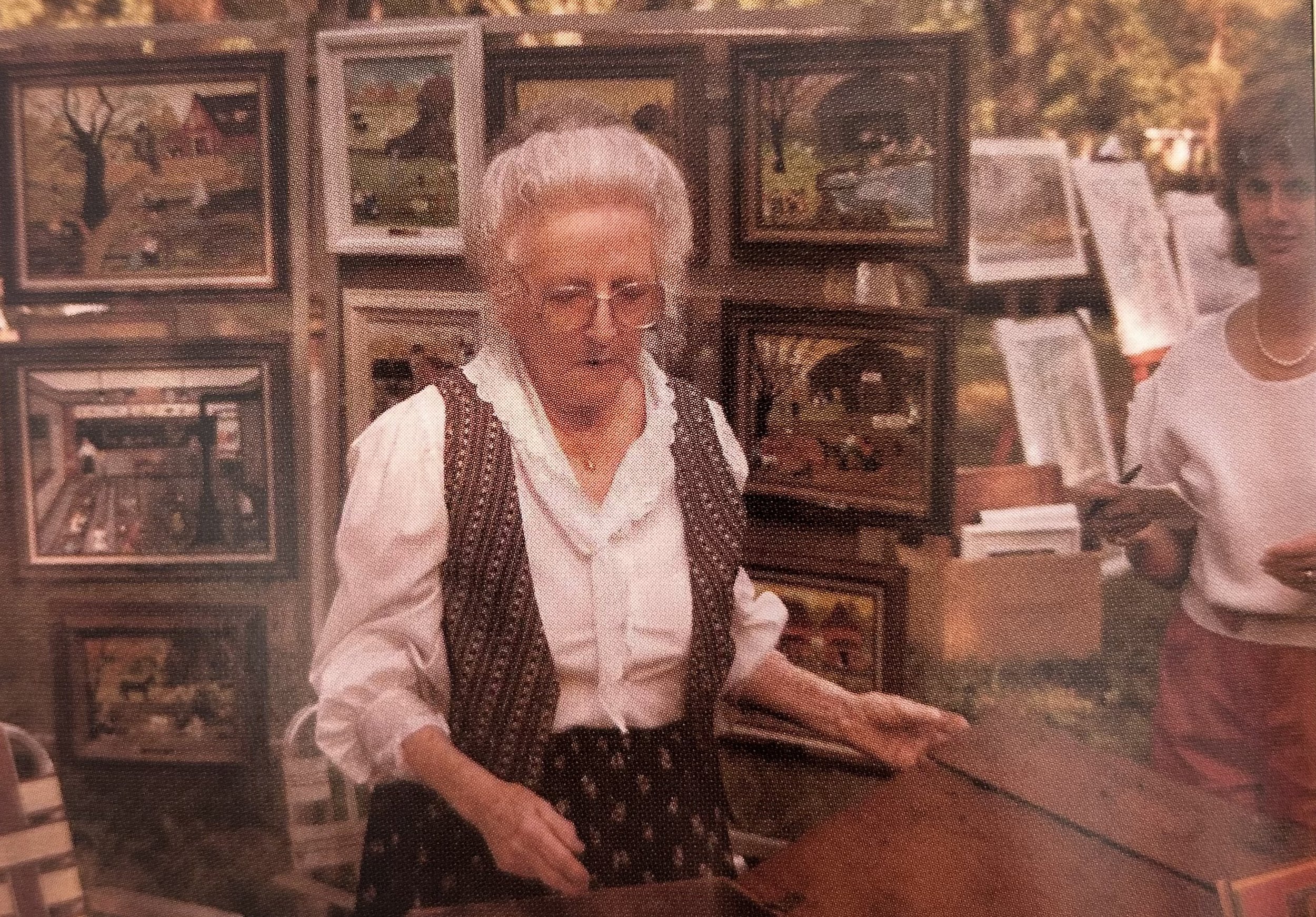 An elderly woman with white hair wearing glasses and a white blouse with a dark vest, standing behind a counter in an art gallery or shop, surrounded by framed paintings and artwork.