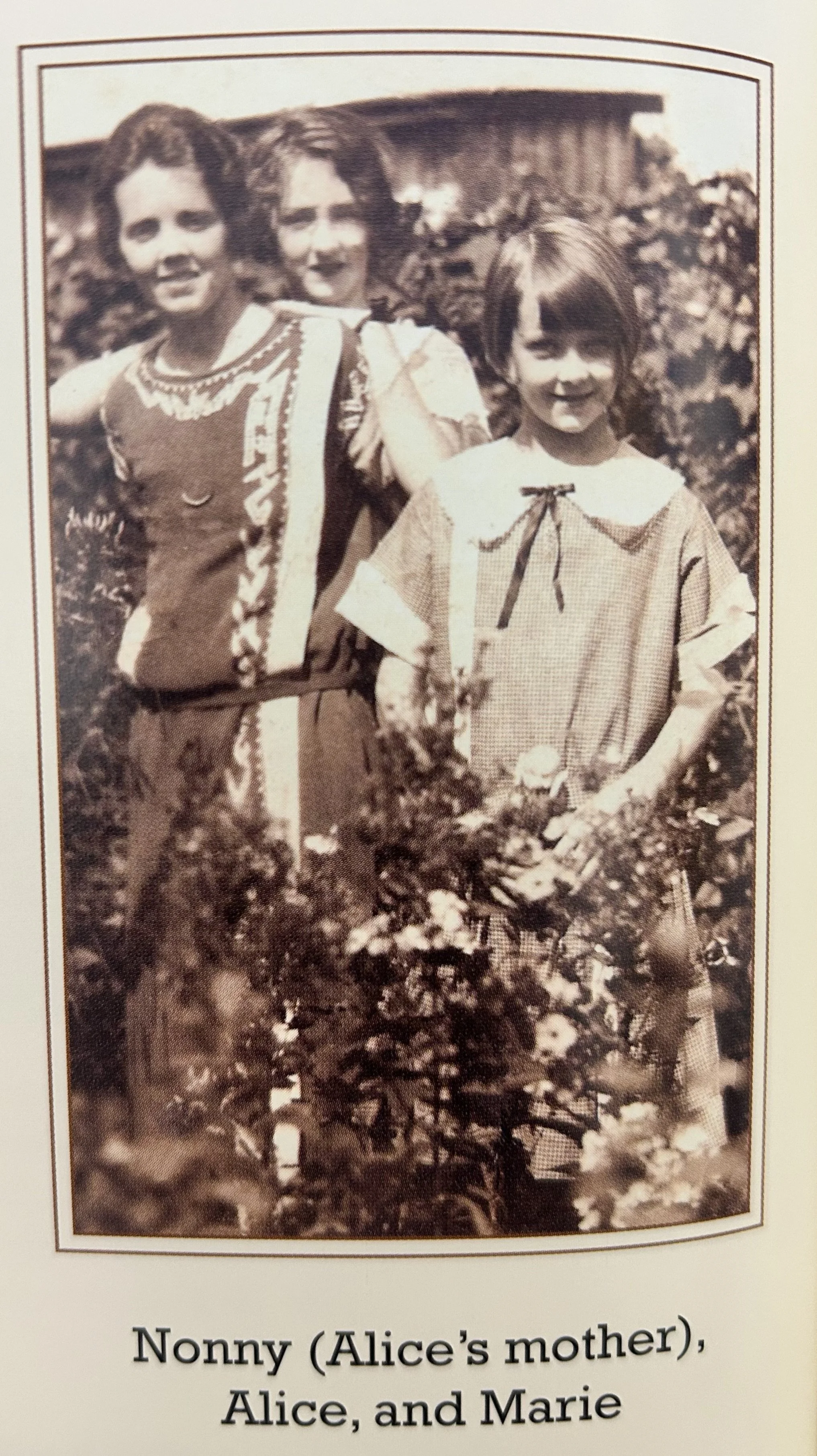A sepia-toned vintage photograph of three women standing outdoors among flowers and foliage, with a wooden building in the background. The woman on the left has short, curly hair and wears a sleeveless patterned dress. The woman in the middle has wavy hair and is smiling, partially obscured behind the other two. The woman on the right has straight hair with bangs and wears a dress with a large collar and a ribbon tie at the neckline.