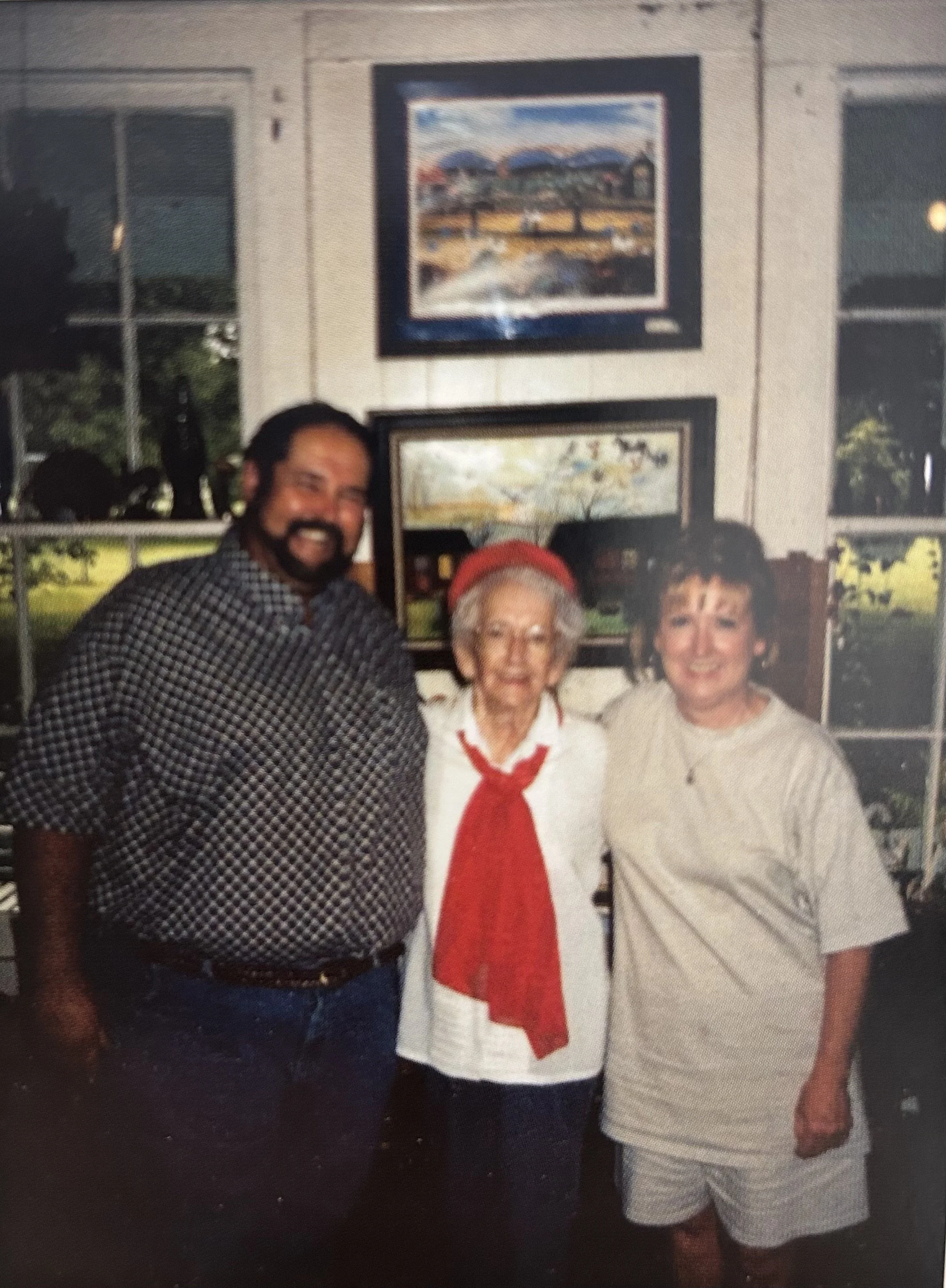 A group of three people posing inside a room with artwork on the wall behind them. The person on the left is a man with a beard, wearing a checkered shirt. The middle person is an elderly woman with glasses, wearing a white shirt with a red neckerchief. The person on the right is a woman with short dark hair, wearing a light-colored shirt.