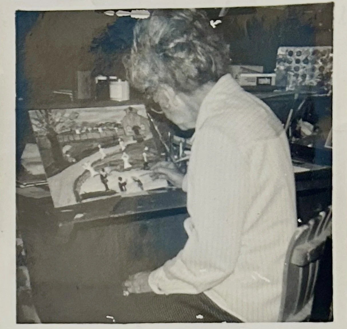 A person with curly hair and glasses is sitting at a desk and working on a painting or drawing of a park scene with trees, pathways, and small figures.