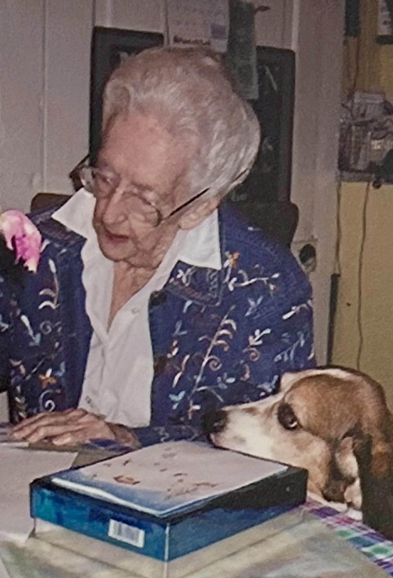 An elderly man with glasses sitting at a table with a dog resting its head nearby. There are some books or boxes on the table in front of him and a TV or monitor behind him.