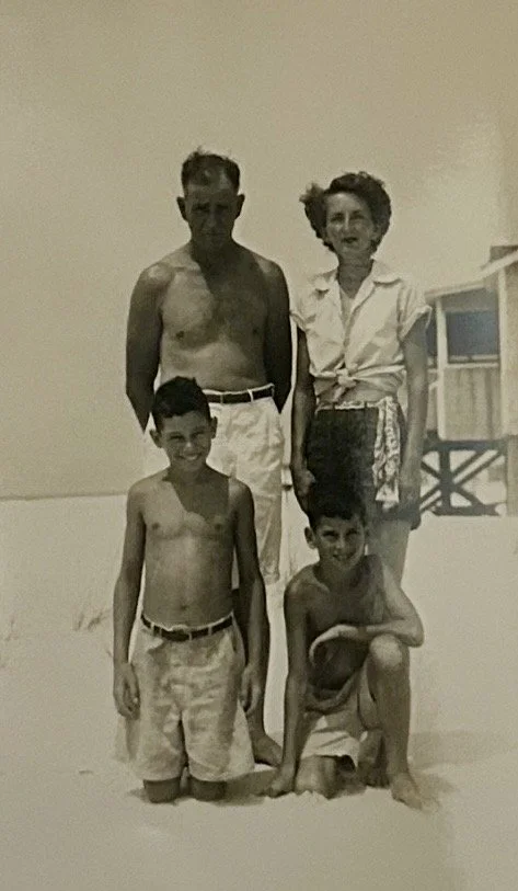 A black and white photo of a family with two adults and two children at the beach. The man is shirtless and wearing light-colored shorts, the woman is wearing a shirt and shorts, the boy standing is shirtless and wearing shorts, and the boy sitting is shirtless and crouching with a hand on his knee.