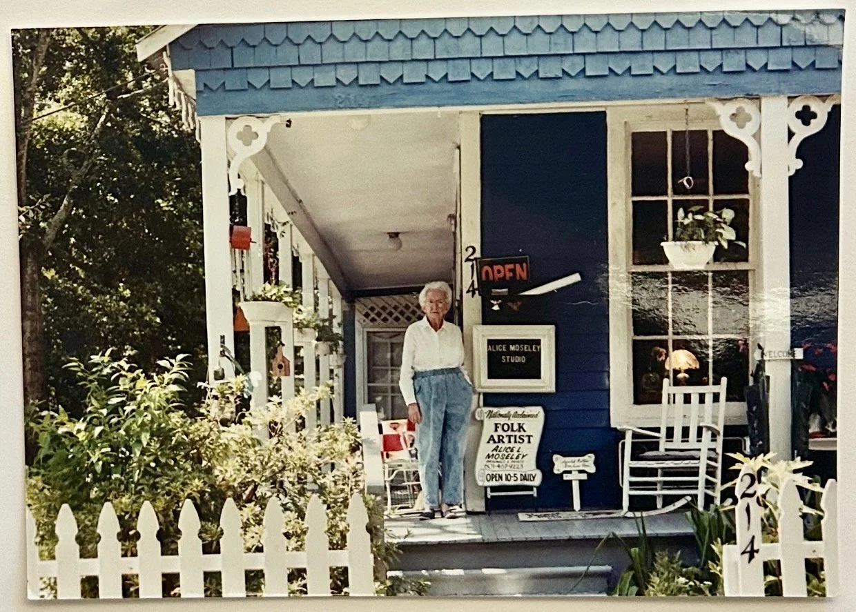 An elderly woman stands on the porch of a blue house with white trim, surrounded by a white picket fence and greenery. The house has signs indicating it is Alice Moseley's studio, a folk artist, and shows the studio's opening hours and welcoming signs.