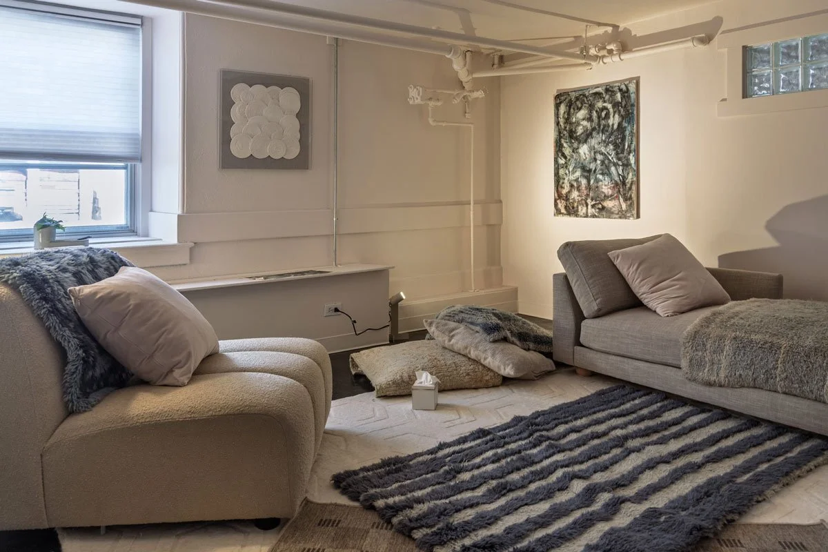 A cozy therapy room with two couches, a window with blinds, abstract art on the walls, pillows, a striped rug, and scattered cushions on the floor in East Falls, Philadelphia.