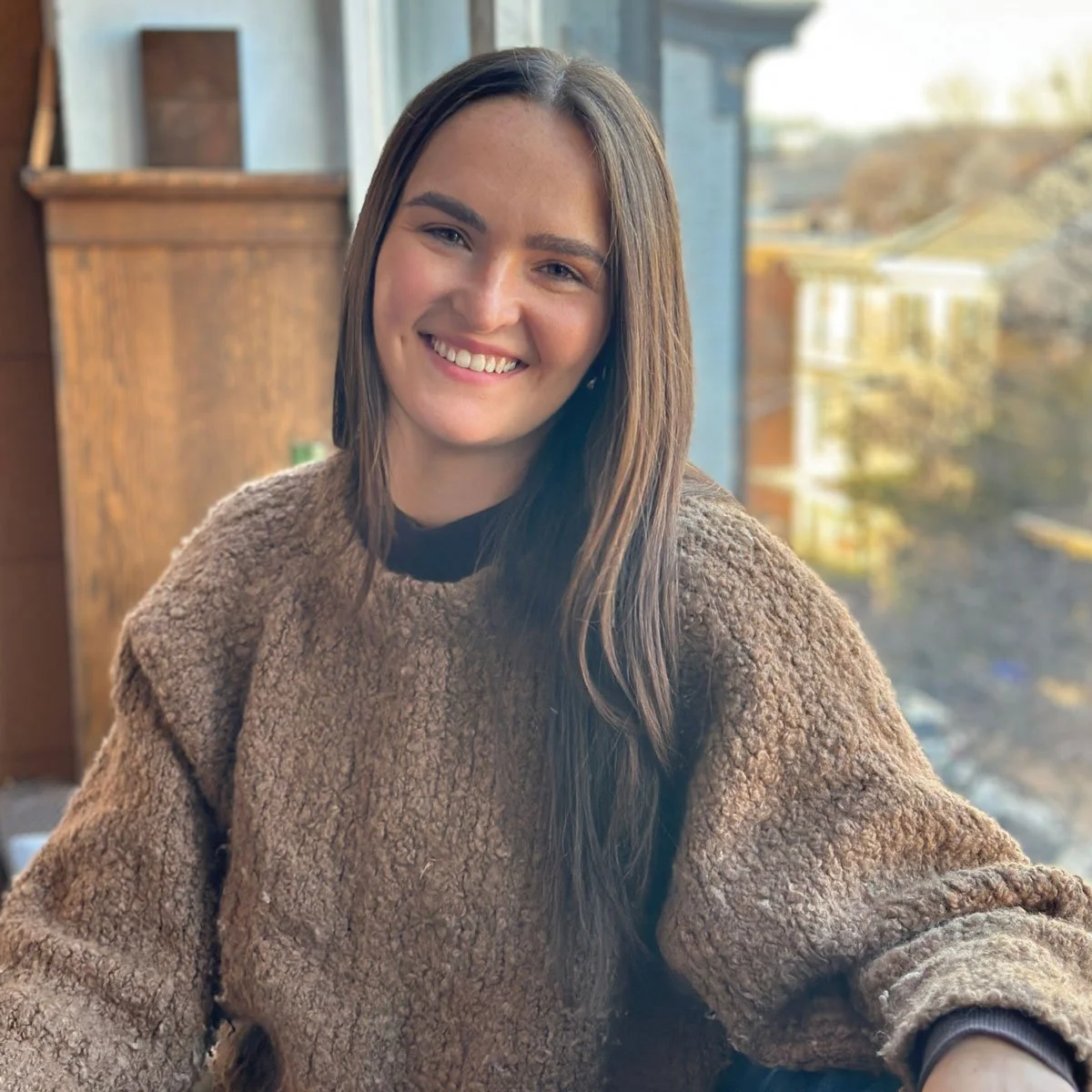 A young woman with long brown hair smiling, indoors near a window with a blurred outdoor scene in East Falls, Philadelphia.