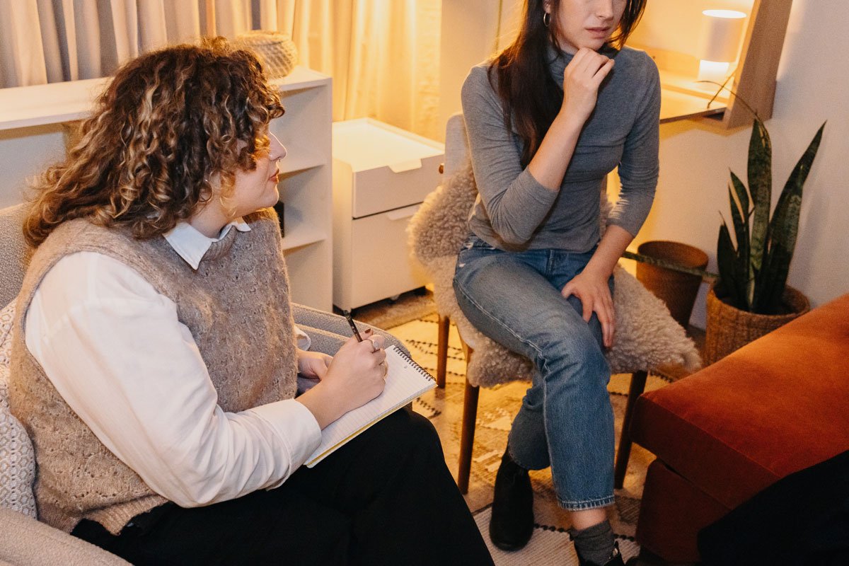 Two women, one with curly hair sitting on a couch taking notes, and the other with straight hair in a gray turtleneck and jeans sitting on a chair with her hand on her chin, in a warmly lit room in East Falls, Philadelphia.