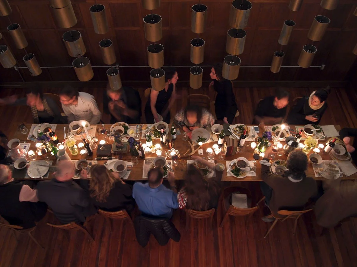 A group of people gathered around a long dinner table set for a meal, decorated with flowers, candles, and wine bottles, in a warmly lit dining room in East Falls, Philadelphia.