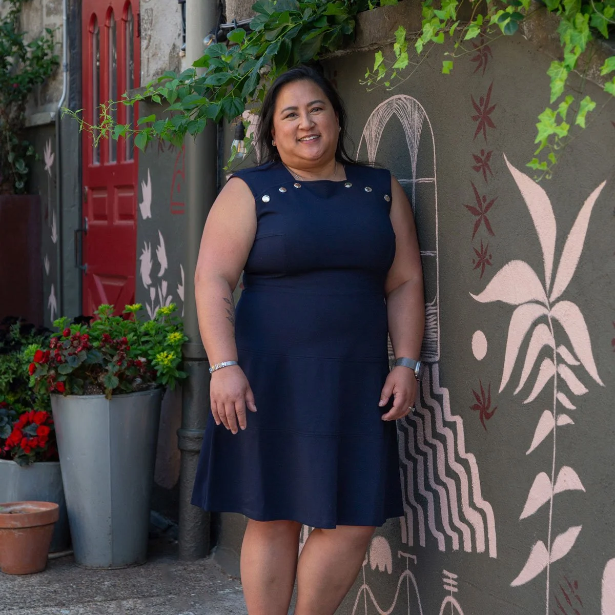 A woman standing next to a wall with abstract flower and fountain art, smiling, wearing a navy blue sleeveless dress with silver button details, surrounded by potted plants and greenery in East Falls, Philadelphia.