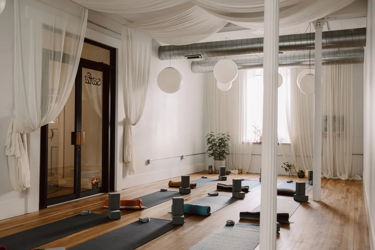 Yoga studio with mats, bolsters, and meditation props arranged on wooden floor, surrounded by sheer white curtains, plants, and soft lighting in East Falls, Philadelphia.