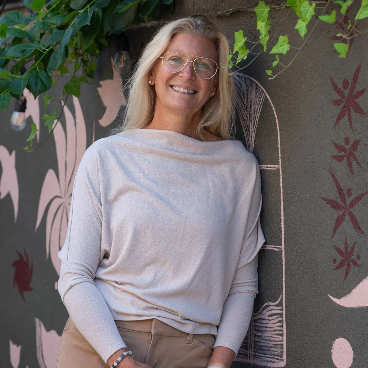 A smiling blonde woman wearing glasses and a white long-sleeve shirt, standing outdoors against a wall with leaf and nature-inspired chalk art, and some green leafy plants hanging nearby in East Falls, Philadelphia.