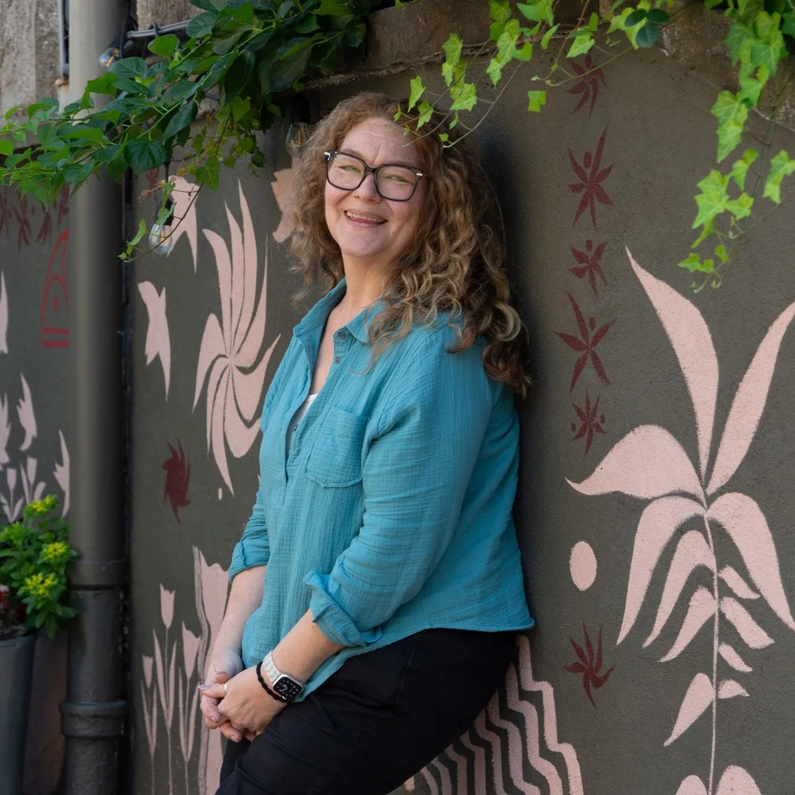 A woman with curly hair, glasses, and a smile, leaning against a wall with pink and red floral and leaf mural, wearing a light blue shirt with rolled-up sleeves and black pants, standing outdoors near green foliage in East Falls, Philadelphia.