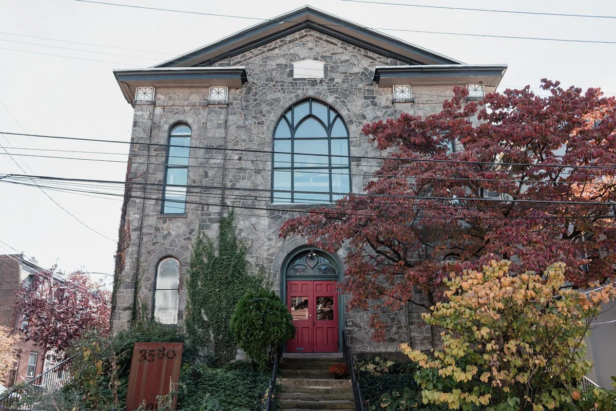 Stone church with arched windows and a red door, surrounded by autumn trees with red and yellow leaves, and ivy growing on its facade in East Falls, Philadelphia.