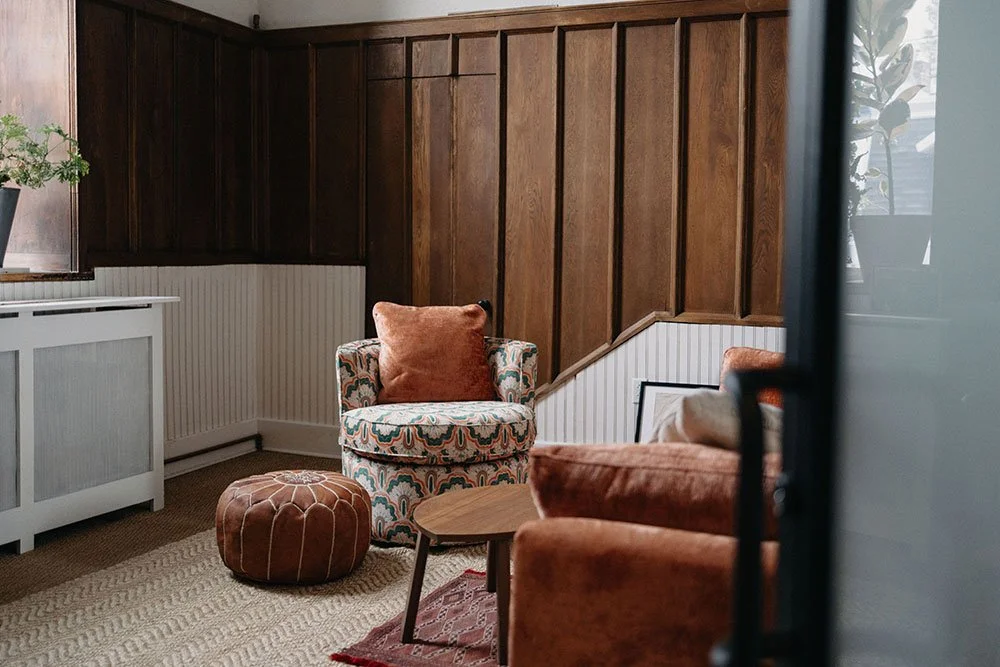 Cozy therapy room in East Falls, Philadelphia with wood-paneled walls, a patterned armchair with a rust-colored cushion, a wooden coffee table, and a burnt orange sofa. There is a window with sunlight and potted plants in East Falls, Philadelphia.