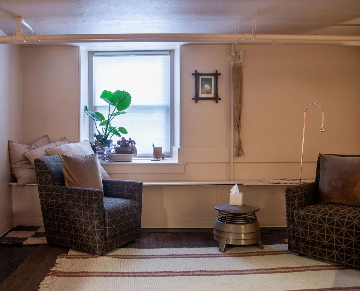 Cozy therapy room with a window, potted plant, armchairs, side table with tissue box, and decorative wall hanging in East Falls, Philadelphia.