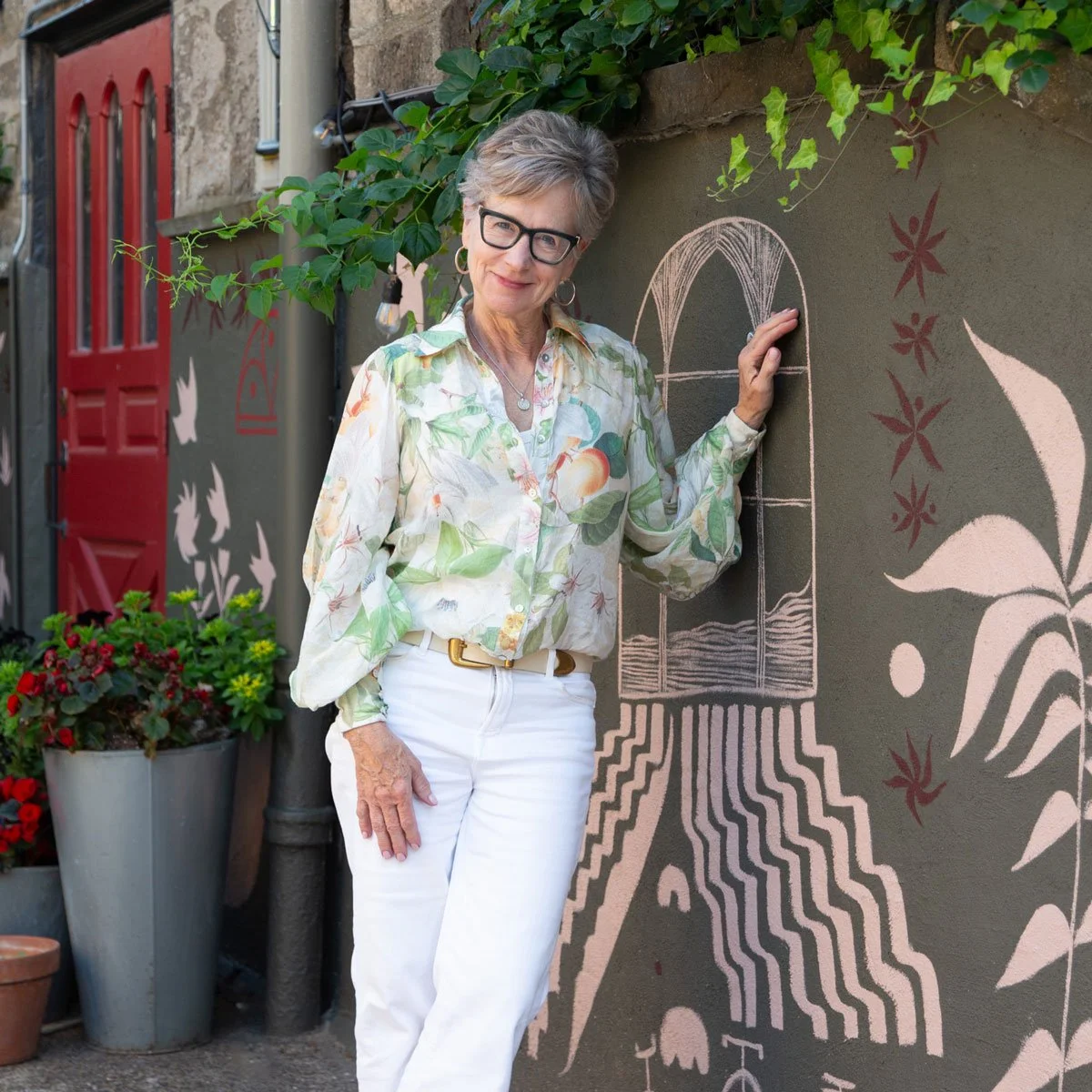 An elderly woman with short grey hair and glasses smiling and standing next to a chalk art mural on a wall. The mural includes a stylized window with flowing lines and floral designs in East Falls, Philadelphia.