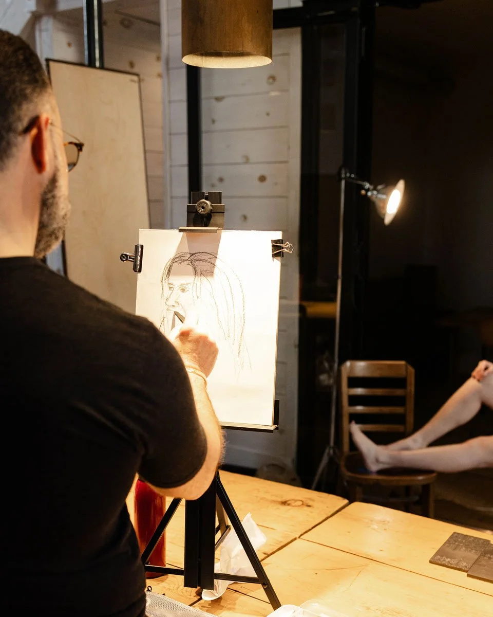 An artist sketching a portrait of a person on a drawing board in a cozy, well-lit room with wooden walls and furniture in East Falls, Philadelphia.
