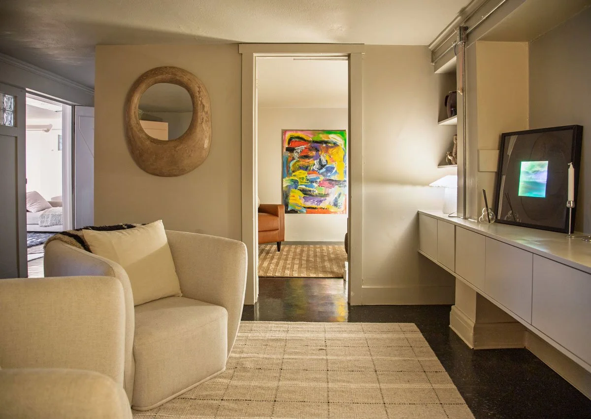 Therapy room with beige armchair, wall mirror, white cabinet with framed picture, colorful abstract painting in the background, and doorway leading to another room with additional artwork and a chair in East Falls, Philadelphia.