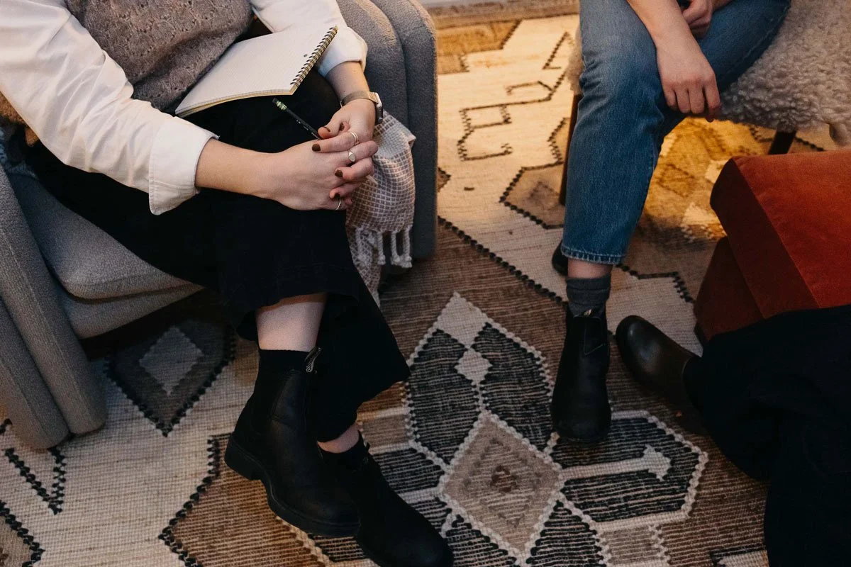 Two people sitting on a sofa and a chair, with their legs visible, on a patterned rug. One person is holding a notebook and pen in their lap. A therapy session happening in East Falls, Philadelphia.
