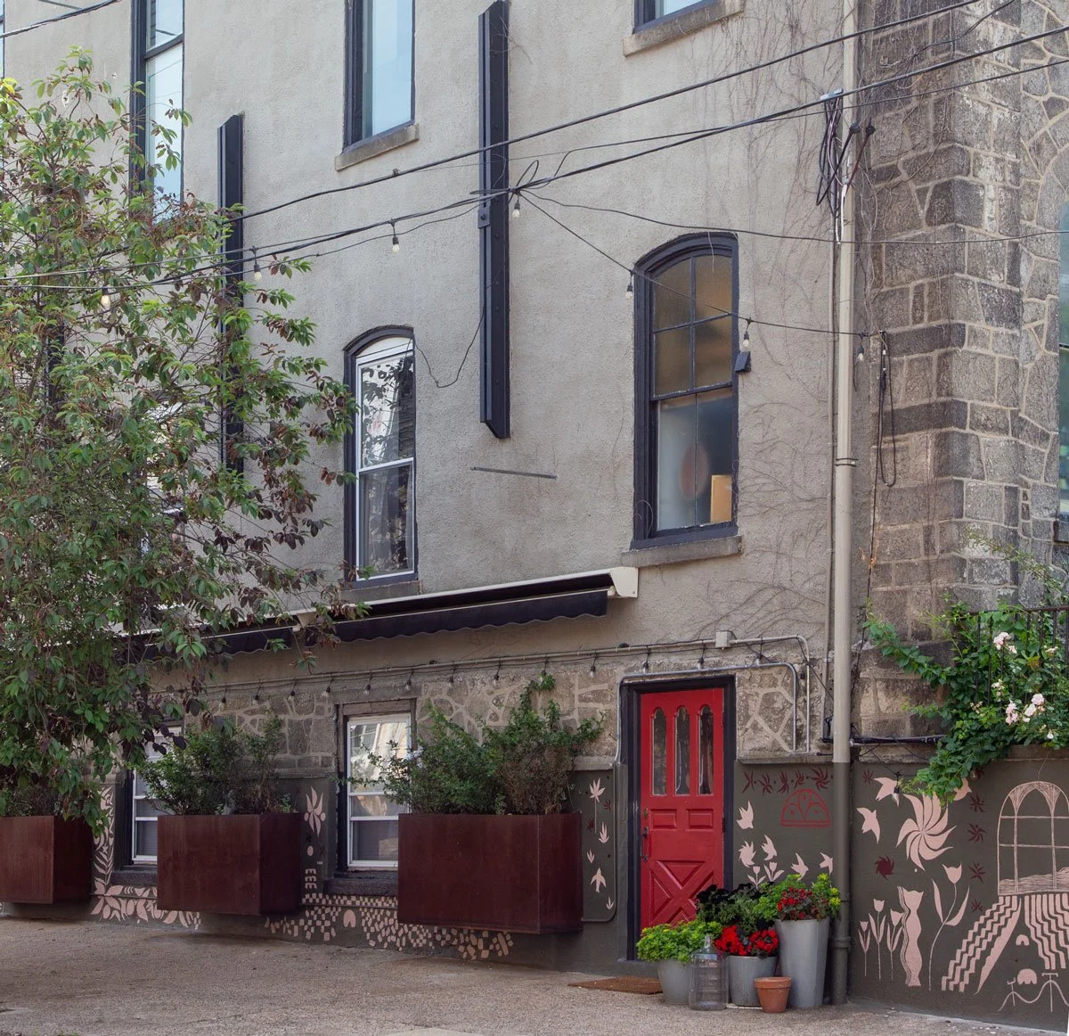 The exterior of a building featuring black-framed windows, decorative black shutters, and a red door. There are potted plants and murals with floral designs and a staircase painted on the gray wall at the ground level in East Falls, Philadelphia.