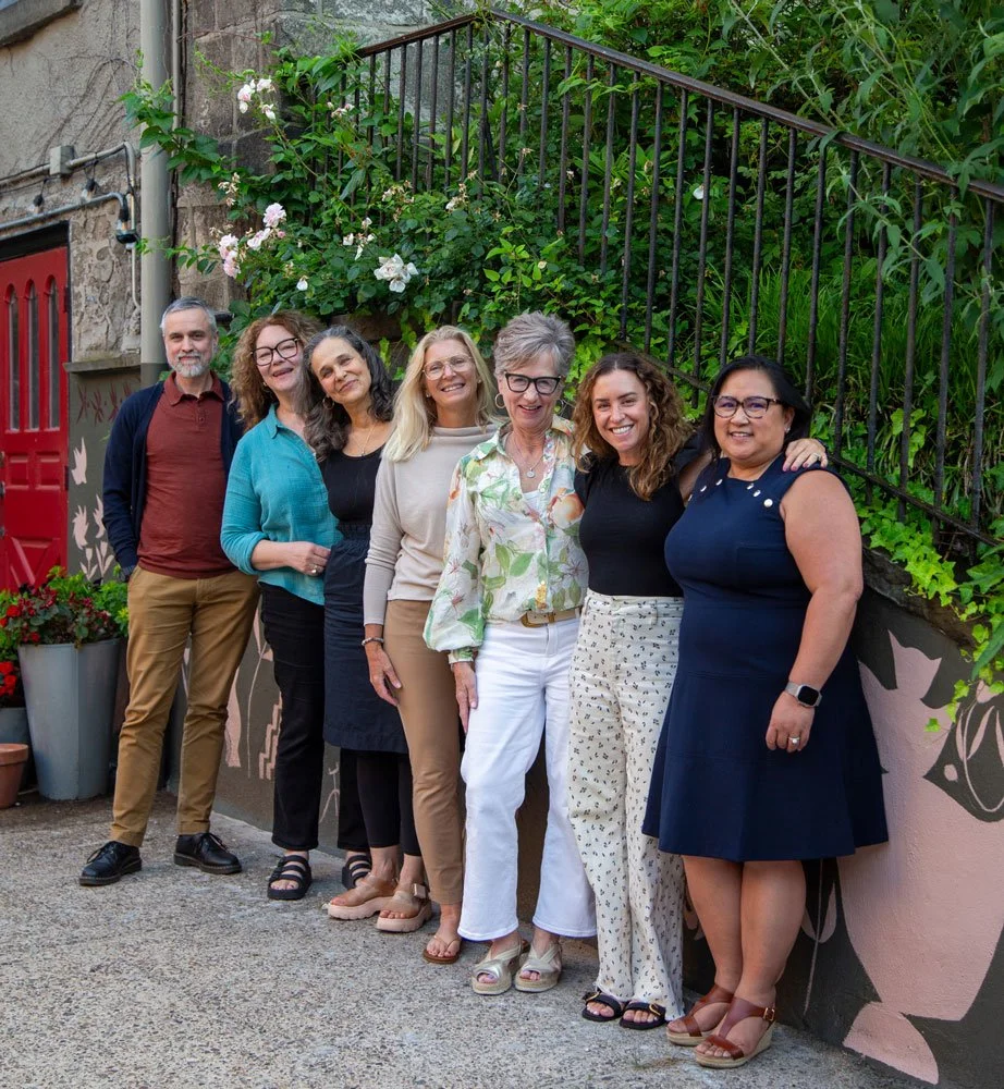 Group of seven women standing outdoors, smiling, in front of a wall decorated with plants and a mural in East Falls, Philadelphia.