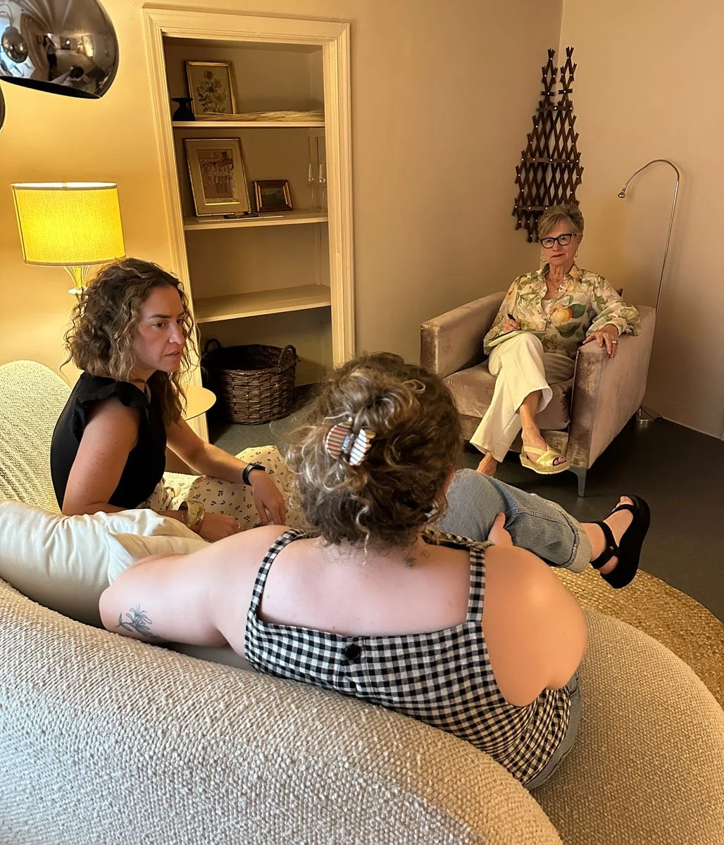 Four women are sitting in a therapy room in East Falls, Philadelphia. The room has warm lighting, with a lamp on a side table, and decorative items on shelves and the wall.